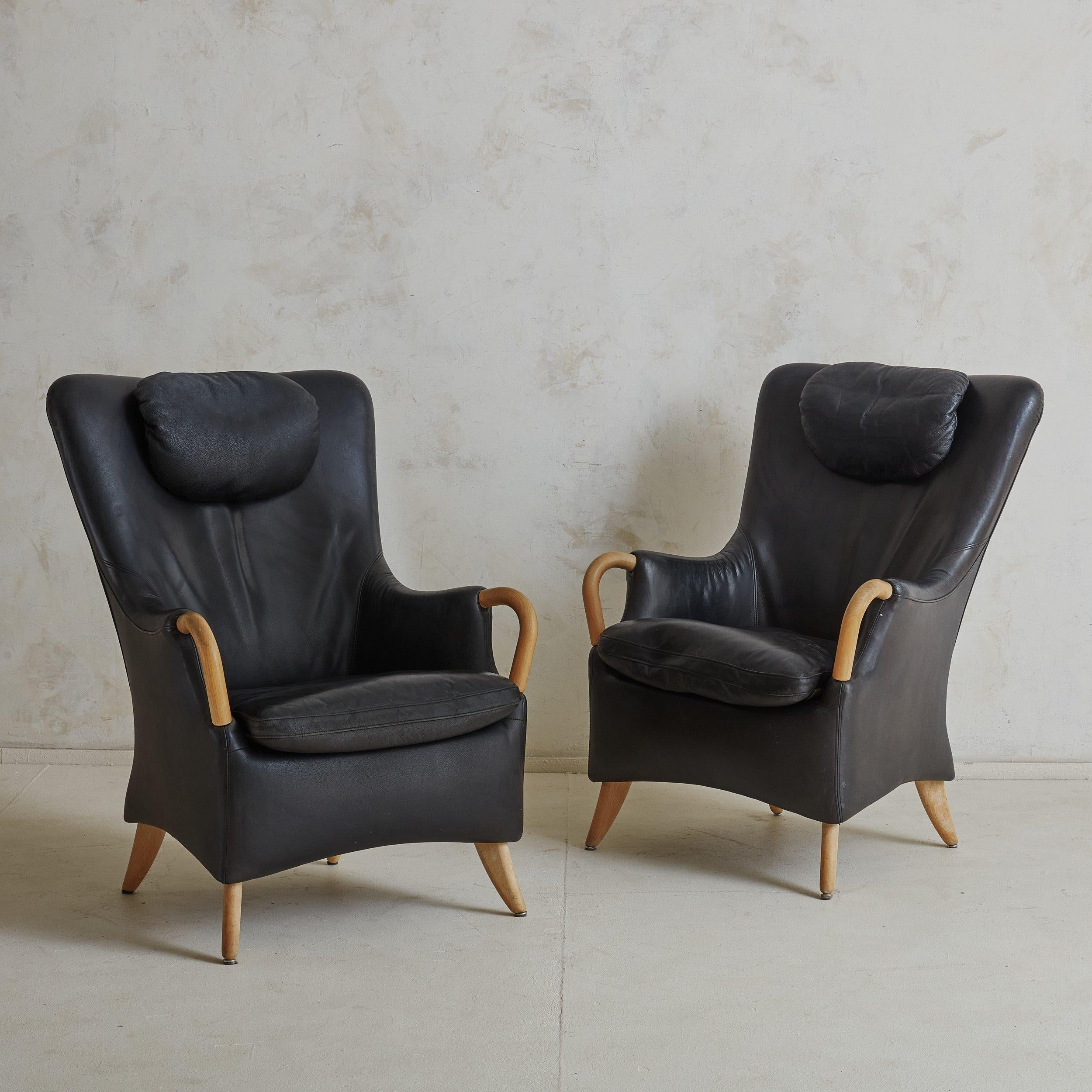 A pair of Danish black leather armchairs with matching footstool designed by Søren Nissen & Ebbe Gehl in 1984. These Scandinavian Modern armchairs feature sculptural, wingback frames and all three pieces are composed of blonde beech wood and