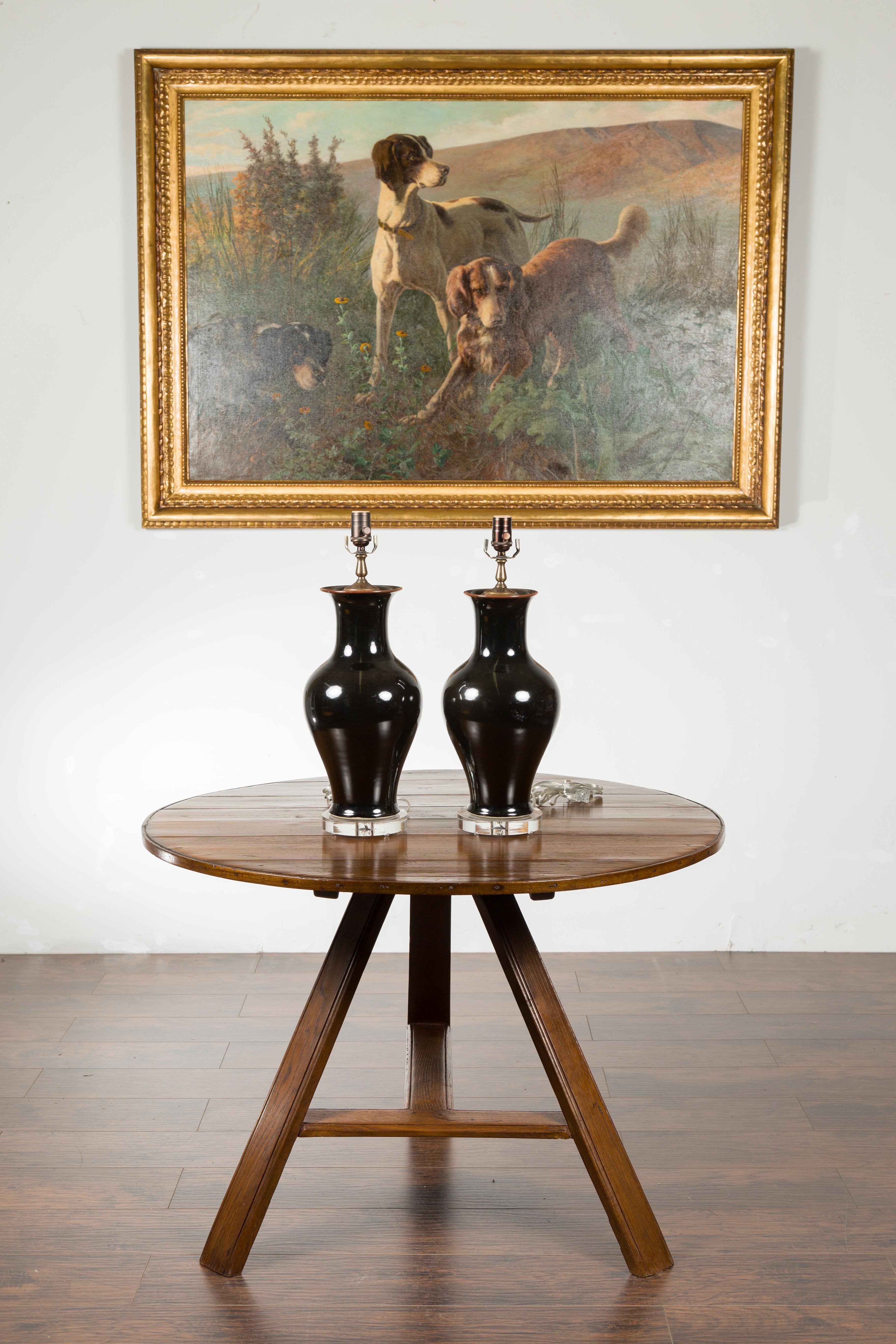 A pair of black porcelain vase-shaped table lamps with lucite bases.  Each of this pair of porcelain table lamps attracts our attention with its sleek lines and black color. Mounted on circular lucite bases and wired for the US, this pair of