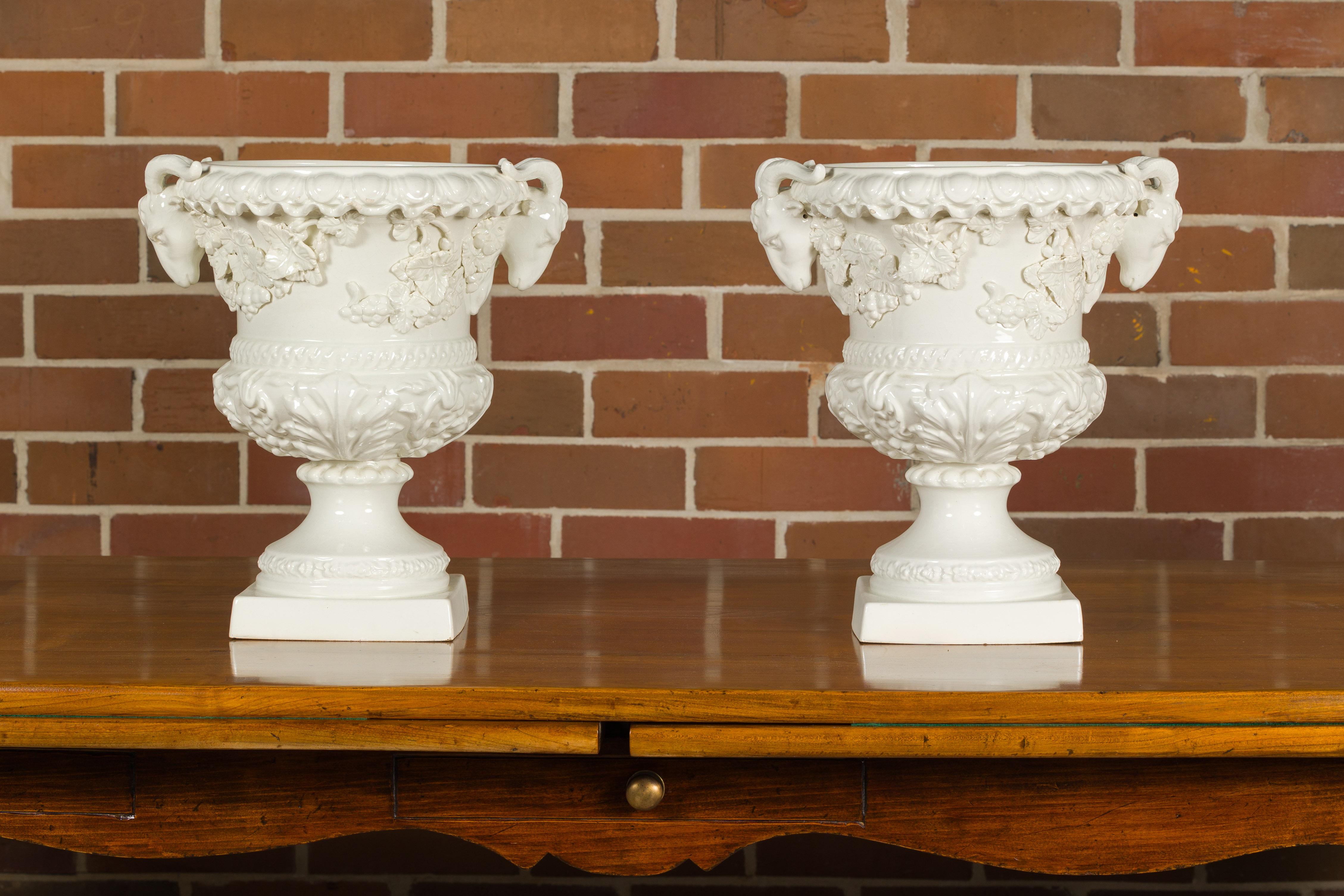A pair of Blanc de Chine urns from the Midcentury period, with rams heads handles, grapes and ivy motifs. This pair of Blanc de Chine urns, originating from the Midcentury period, encapsulates a blend of classical elegance and intricate design. Each