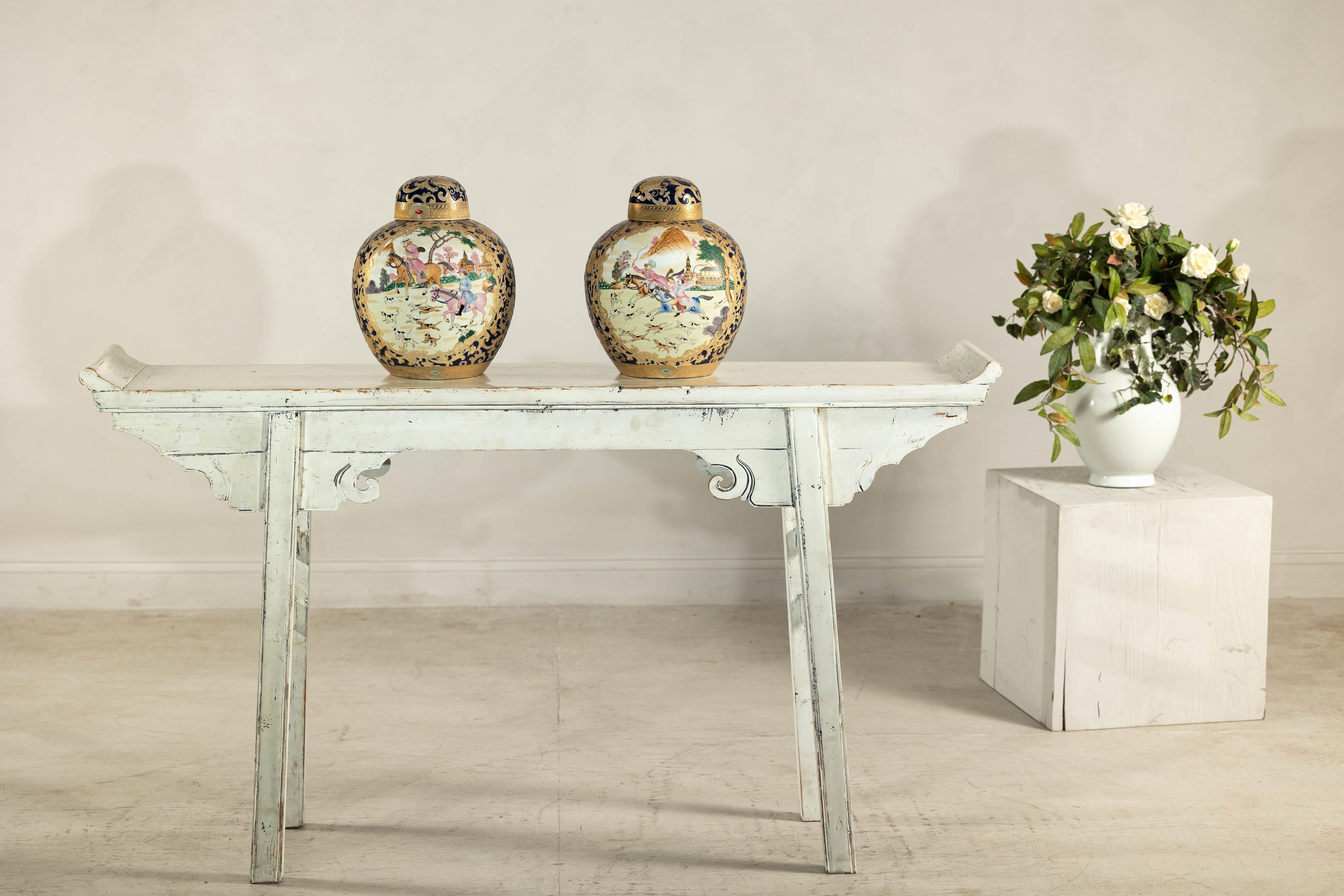 Ricca di opulenza e narrazione, questa coppia di urne con coperchio vintage proveniente dalla Cina presenta un vivido tableau di eleganza equestre. Su un fondo d'avorio lucido, ogni recipiente arrotondato è ornato da scene di caccia dipinte a mano,