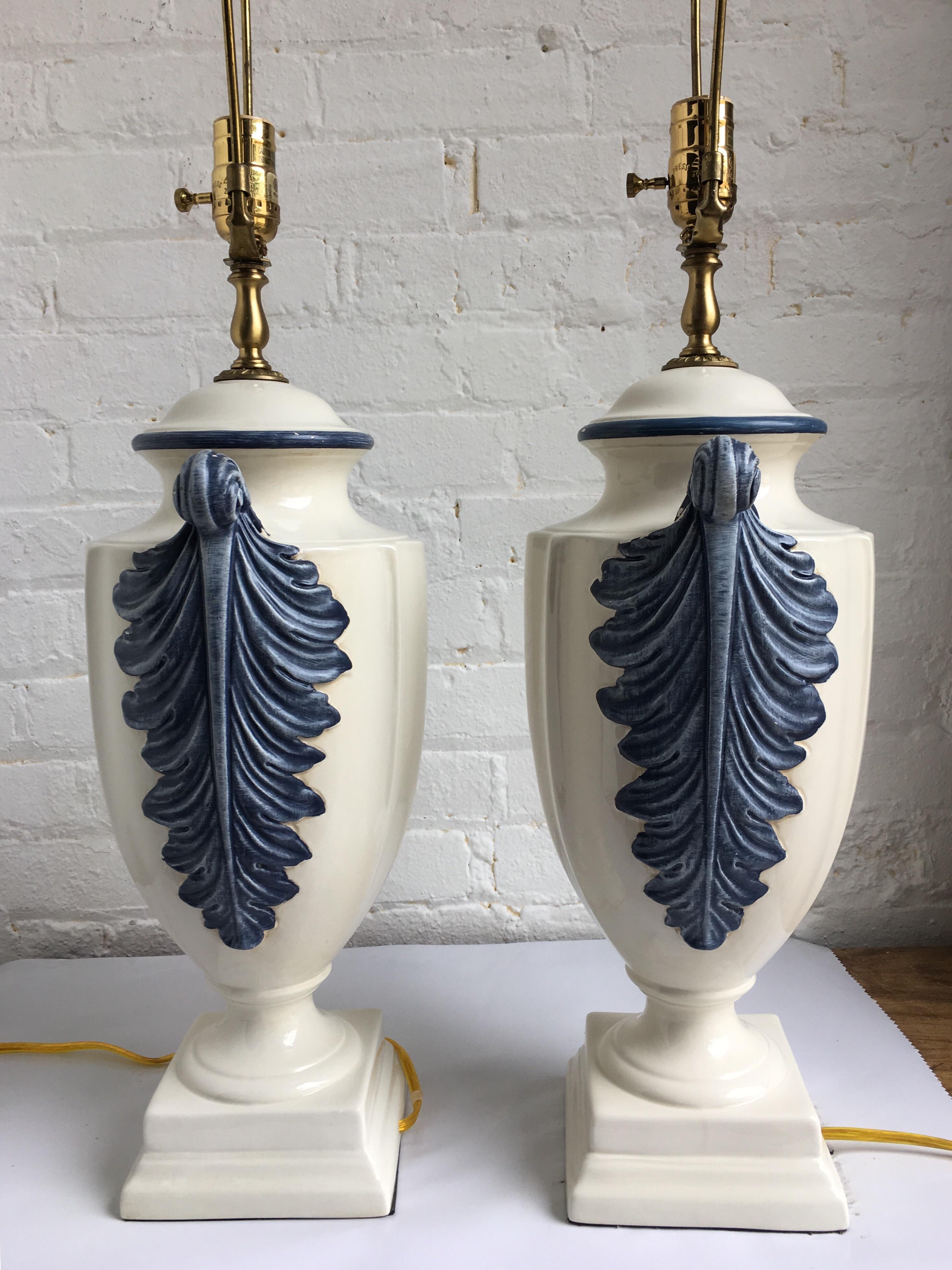 Pair of Hollywood Regency style blue and white porcelain glazed urn table lamps on plinth bases. Side handles feature dimensional hand painted plume or feather decorations. Lamp shades not included. 

Measures: 31.5 inches high to harp.
22.5 inches