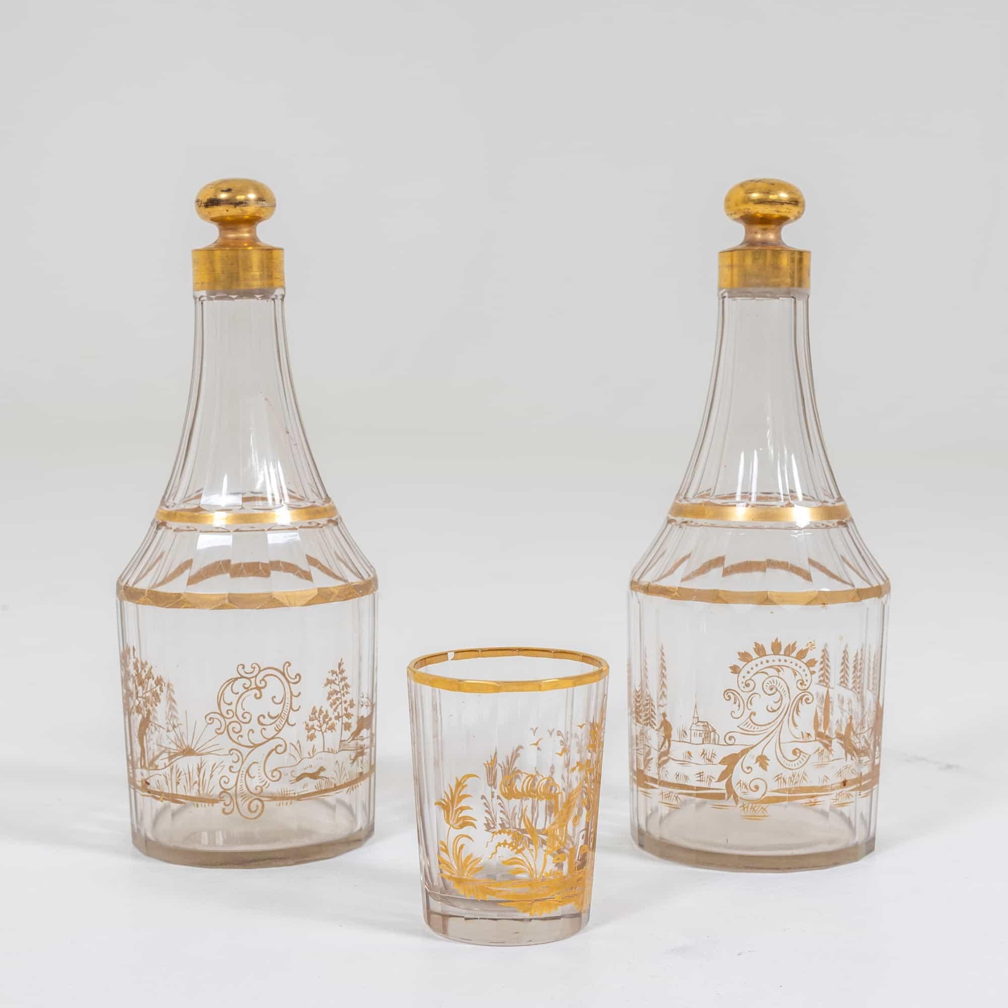 Two faceted carafes made of clear glass with stopper and gold painting. Comes with a matching drinking cup (9.5 x Ø7 cm).