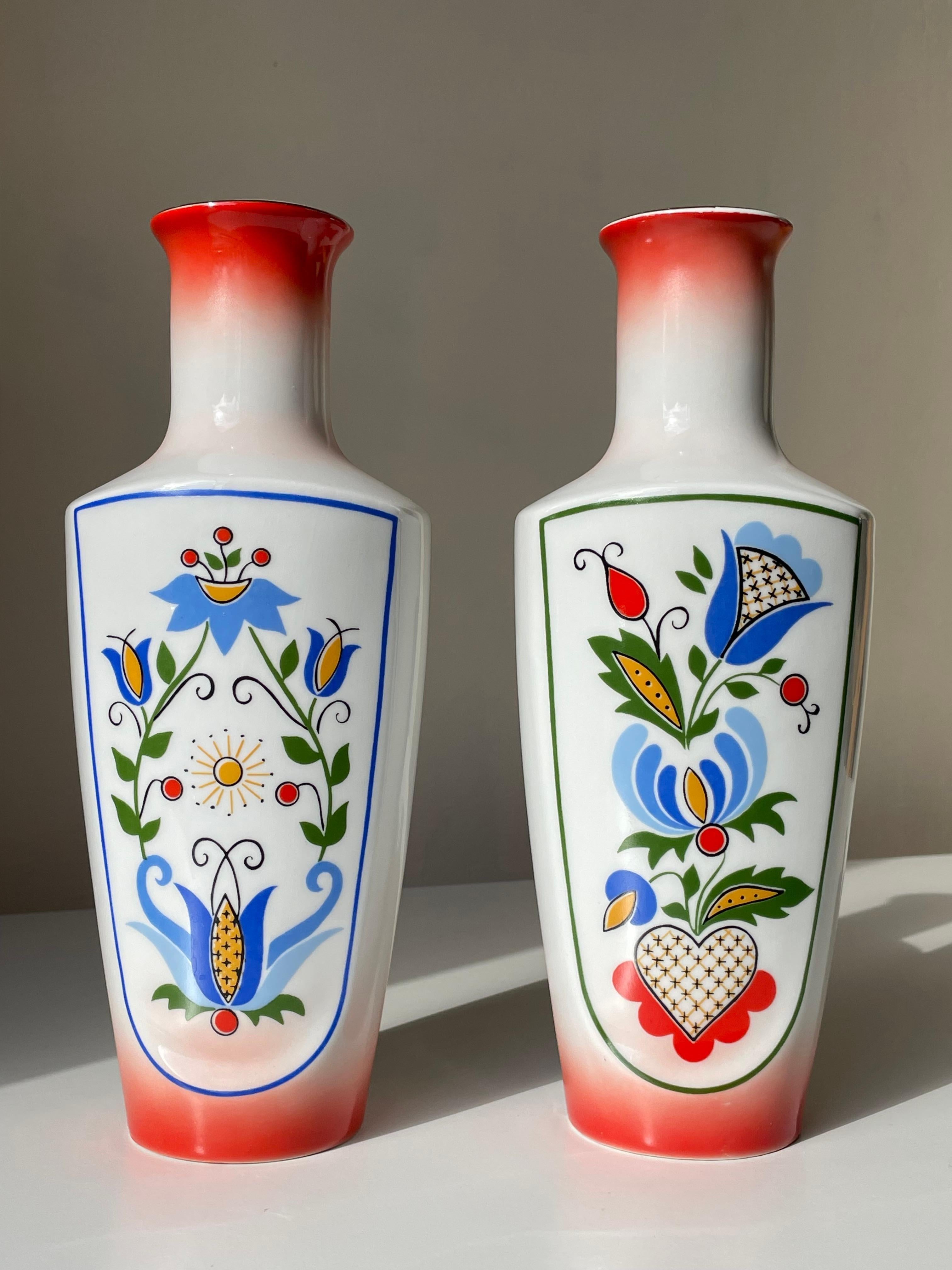 Set of two colorful bohemian 1970s porcelain vases decorated by hand in bright colors. Stylized floral folk art decorations in green, red, yellow and blue colors on white background with bright red base and top with golden lines. Manufactured by