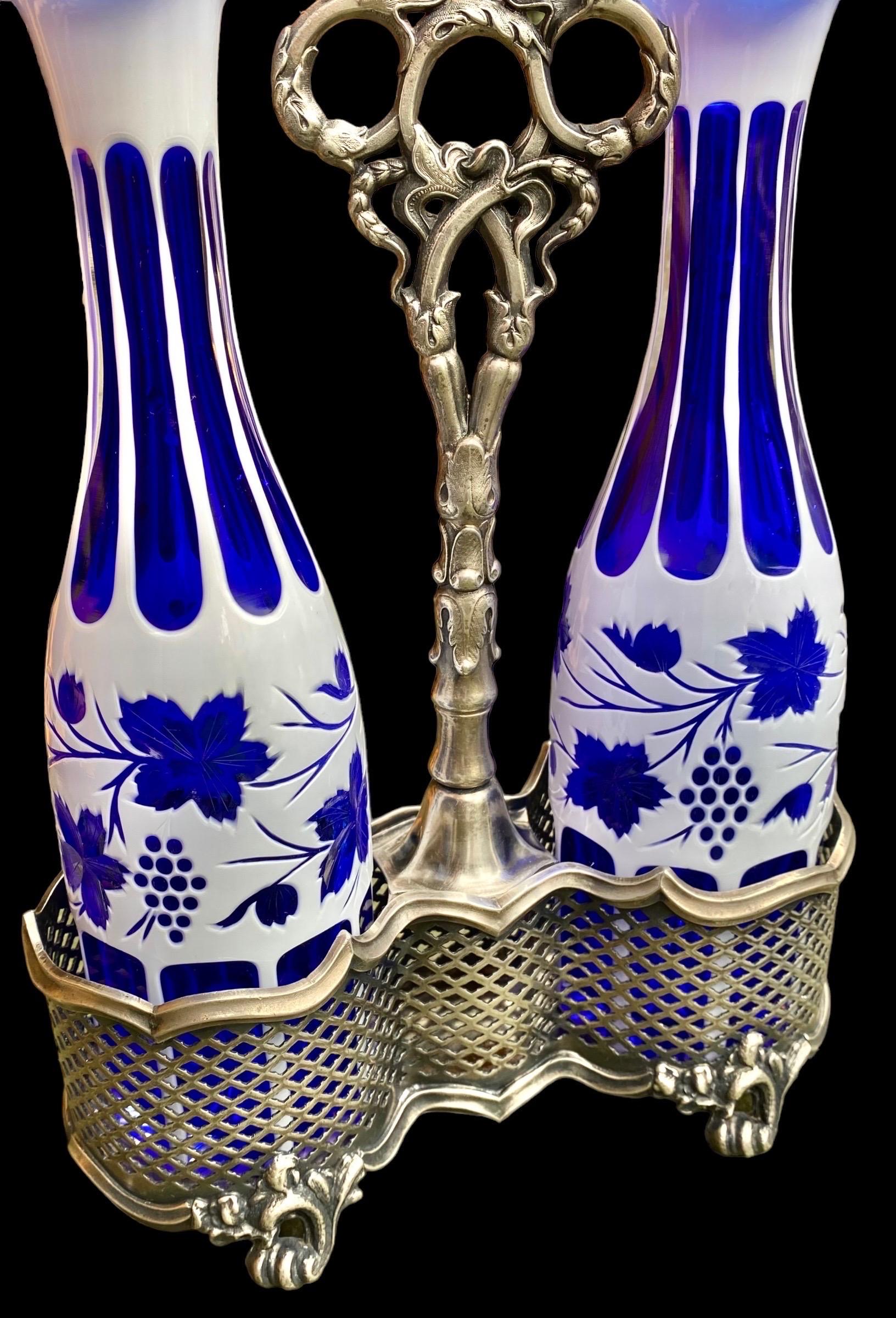 A fine pair of Bohemian glass white over cobalt blue decanters, late 19th century, decorated with cut panels and grapes, set with matching stoppers in a lovely Britannia pierced metal holder with a pierced flower-molded handle and stem.
A delight