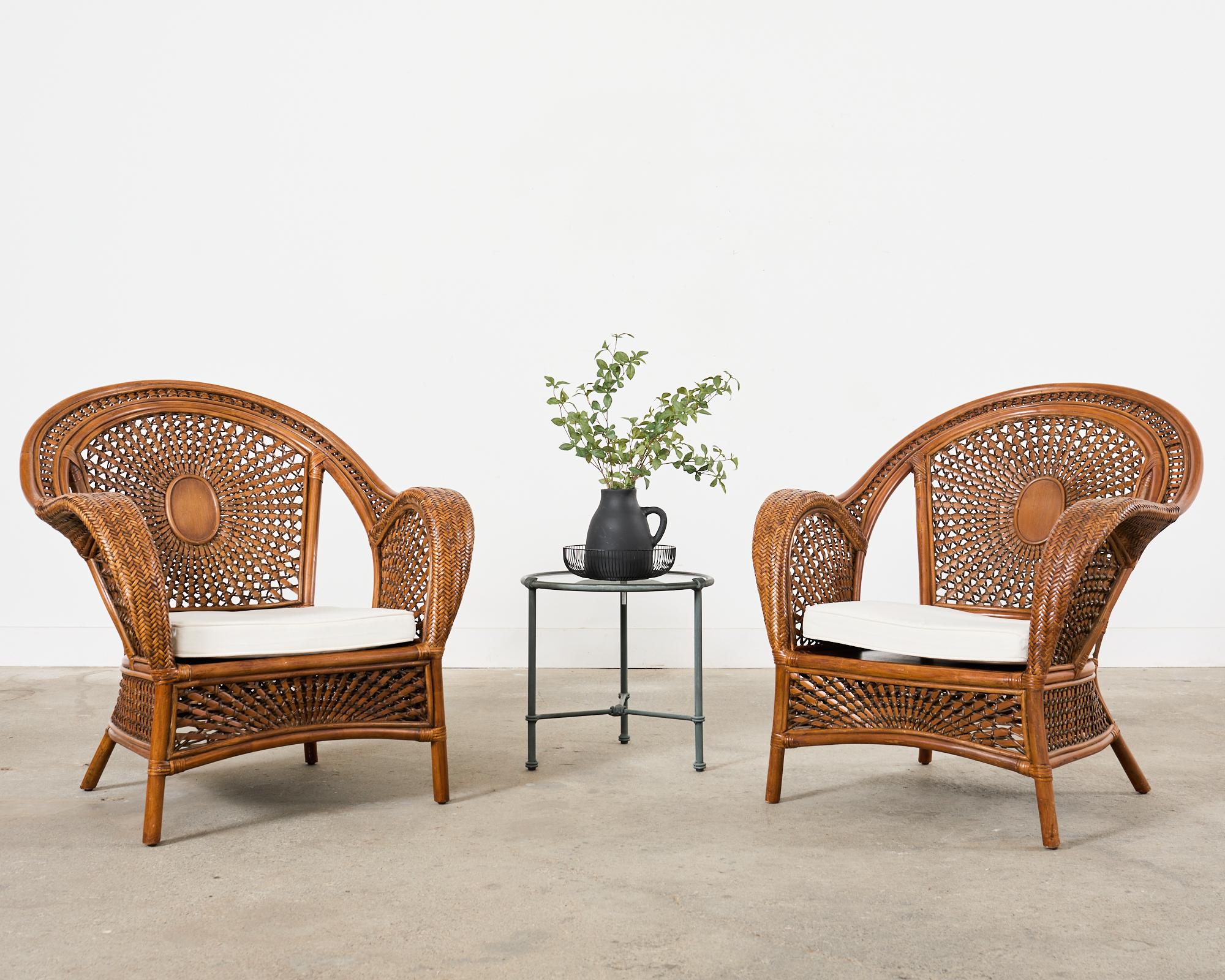 Stylish pair of Bohemian rattan and wicker lounge chairs or armchairs having large peacock or fan backs. The chairs feature gracefully curved rattan frames embellished with decorative woven wicker designs of sunburst patterns. The backs are centered