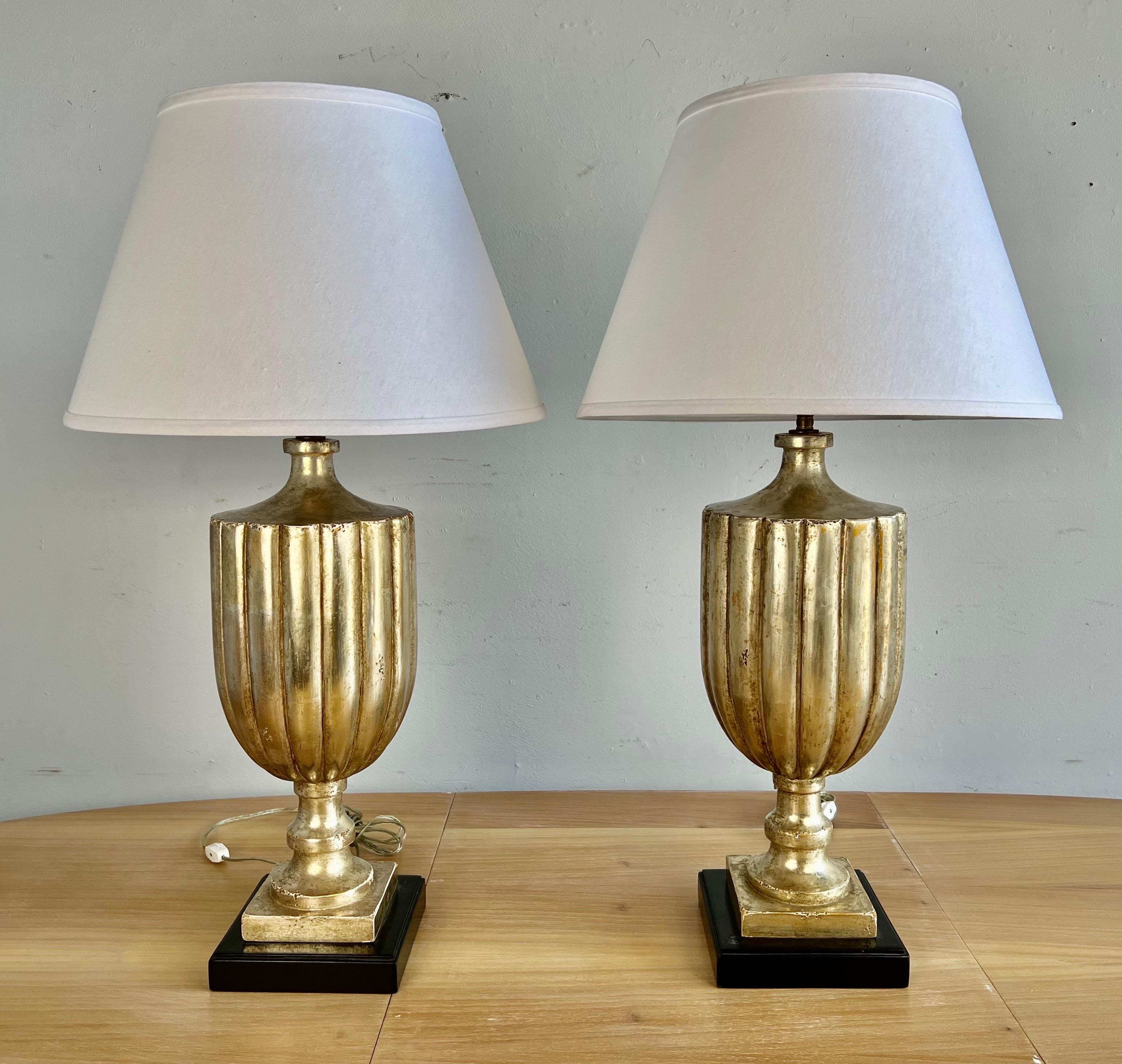 Pair of carved wood fluted urn shaped lamps in a silvery gold finish. The lamps are crowned with white linen shades. Newly rewired and in working condition.