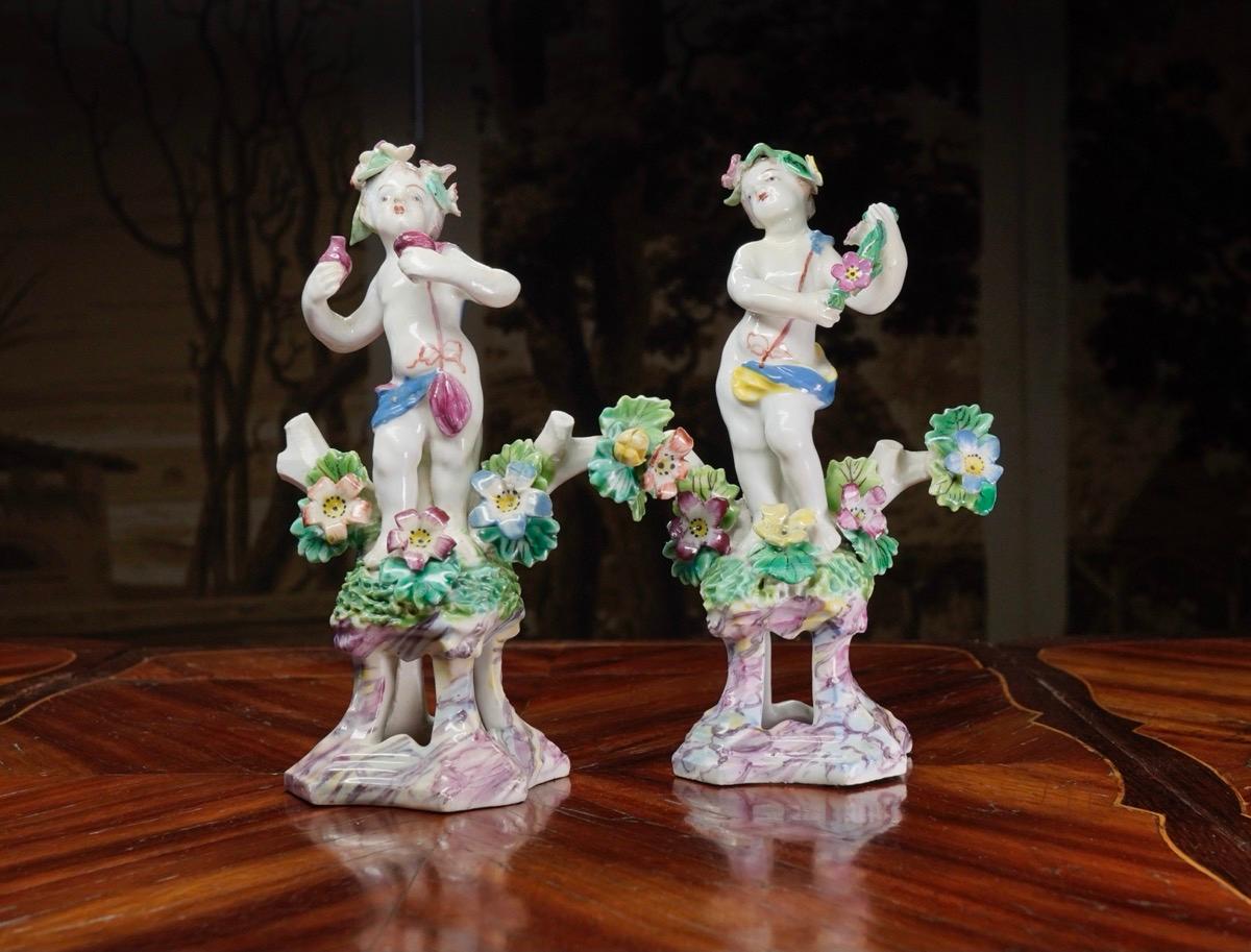 Pair of bow ‘cherubs’, standing cloth draped with a pink ribbon fastening, their hair decked with a flower garland, a flowery bocage sprouting from their grassy mound base, supported on high scroll plinths with cut-out centers, finished in a