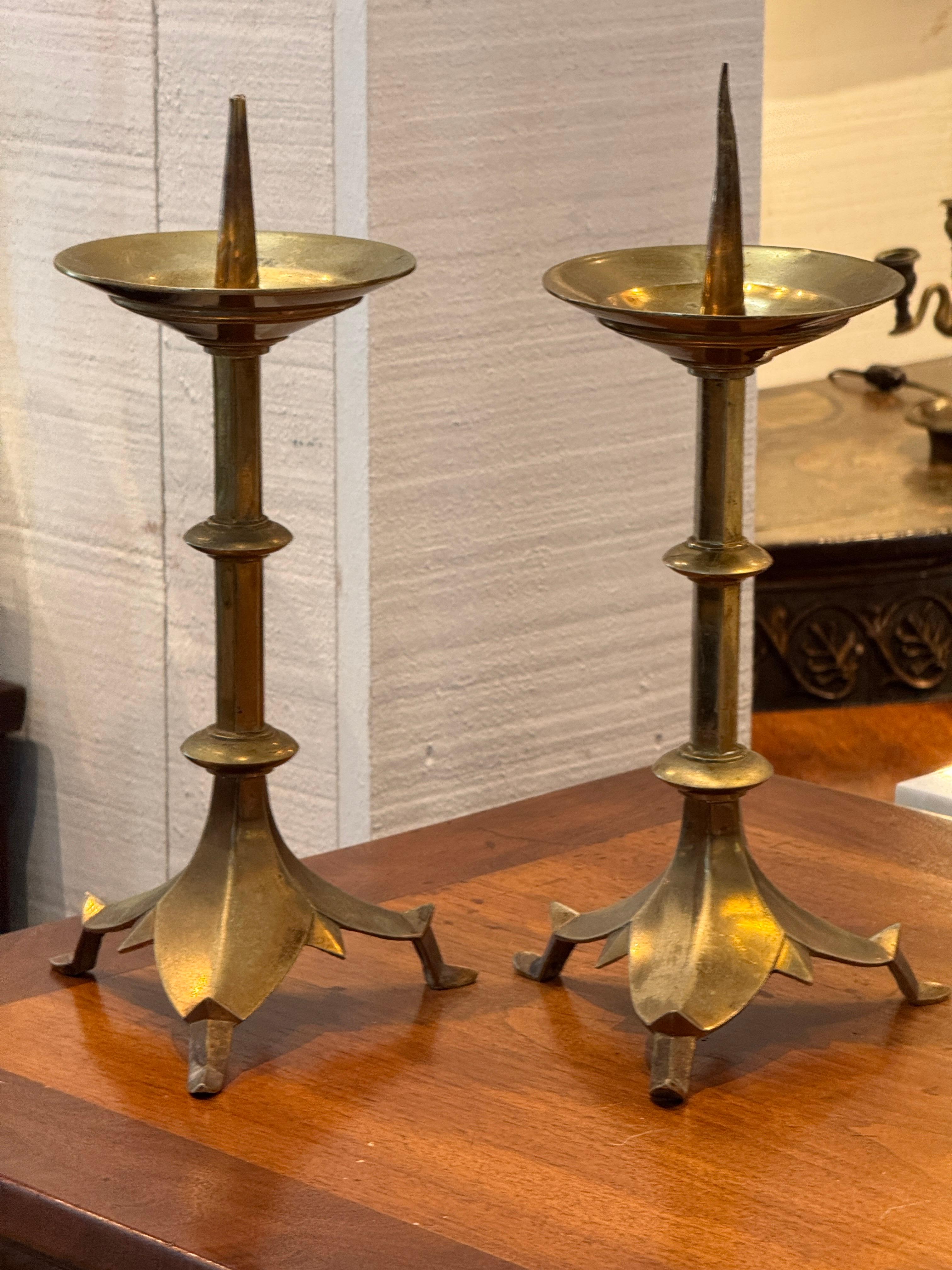 Coppia di bastoni da altare in ottone con colonne alte e affusolate e basi a tripode a forma di petali. Le leccarde sono dotate di sottili punte per fissare le candele. Il loro design equilibrato e la calda patina invecchiata riflettono