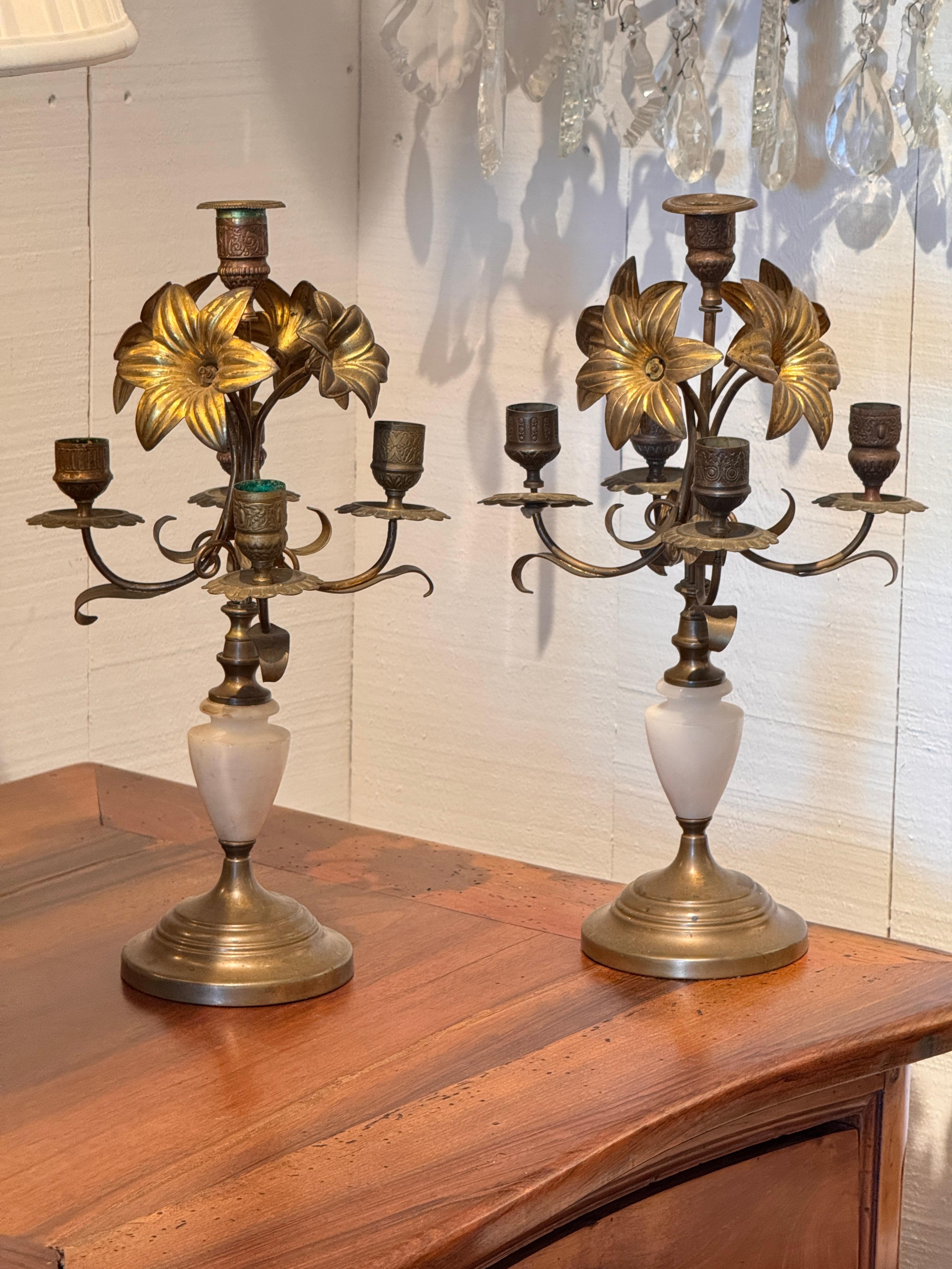 Pair of brass five-arm candelabra featuring lily-shaped floral elements and etched cups. The curving arms rise from turned alabaster bases. Slight verdigris and wear consistent with age.