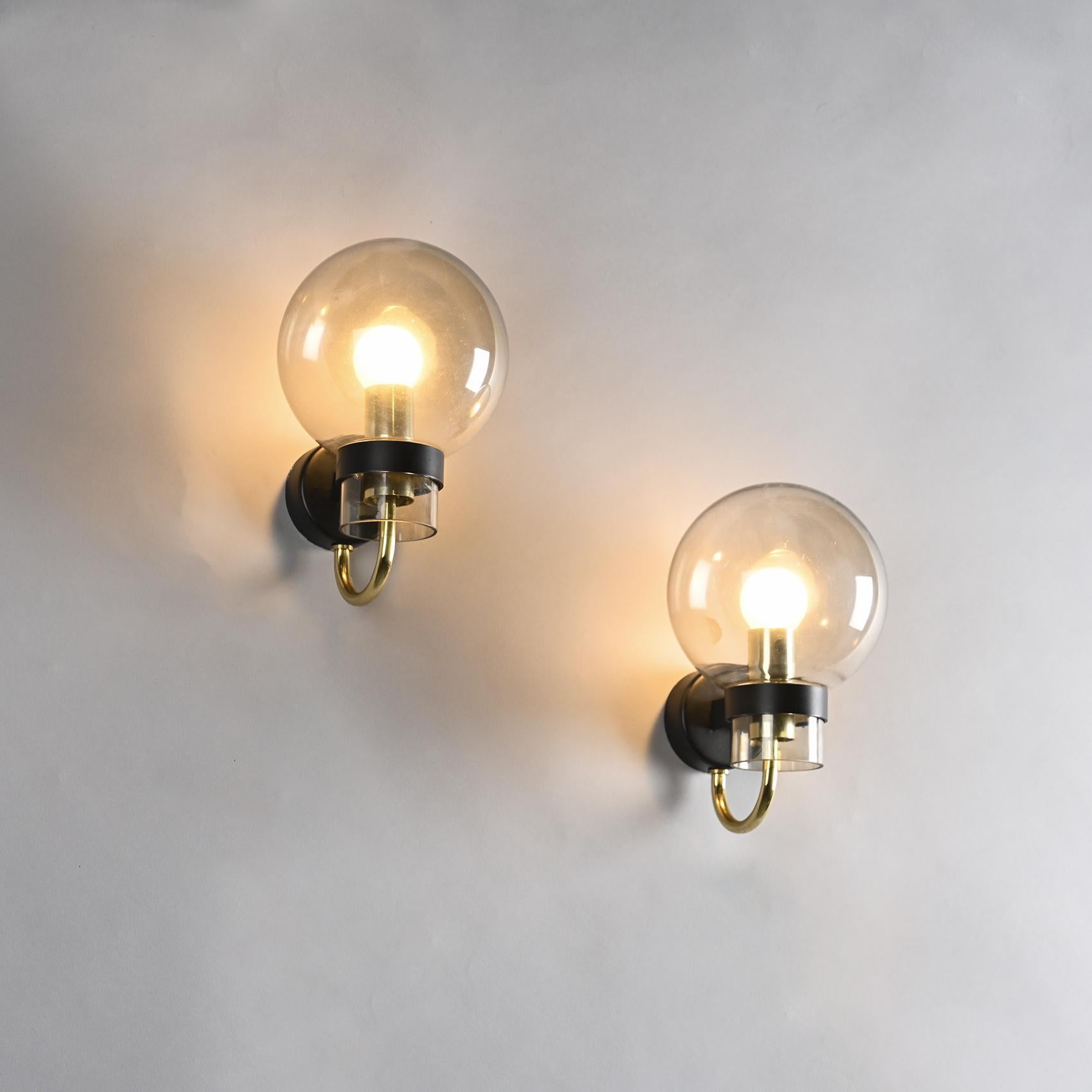 Pair of wall sconces published by Glashütte Limburg, circa 1970.

Each sconce features a black lacquered metal backplate and a single gilt brass arm holding a transparent, subtly iridescent hand-blown glass globe. The composition exudes a discreet