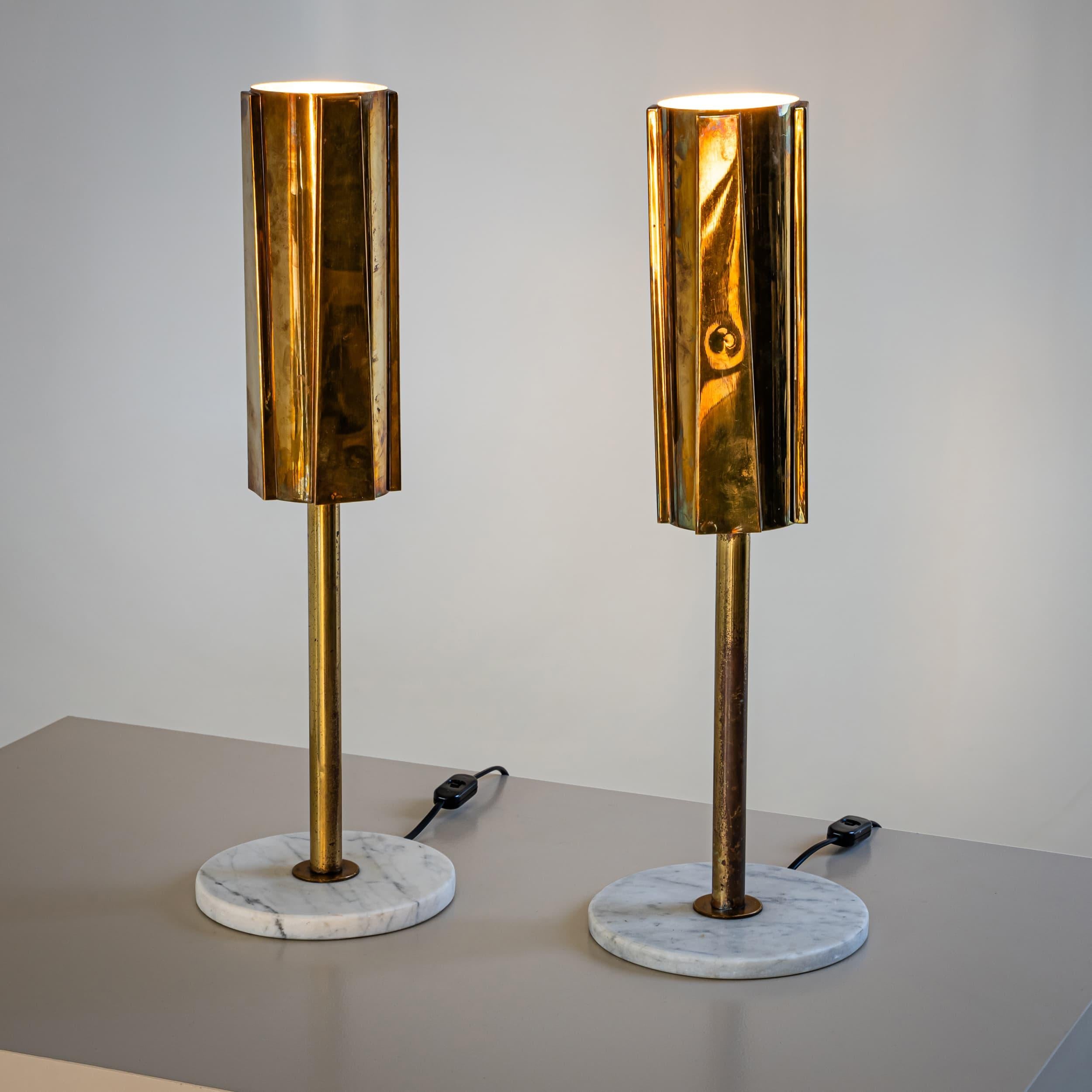 Pareja de lámparas de sobremesa sobre bases circulares de mármol con pantallas cónicas de latón adornadas con elementos trapezoidales aplicados. Cada lámpara está equipada con un único casquillo y emite una luz suave, indirecta y atmosférica. 