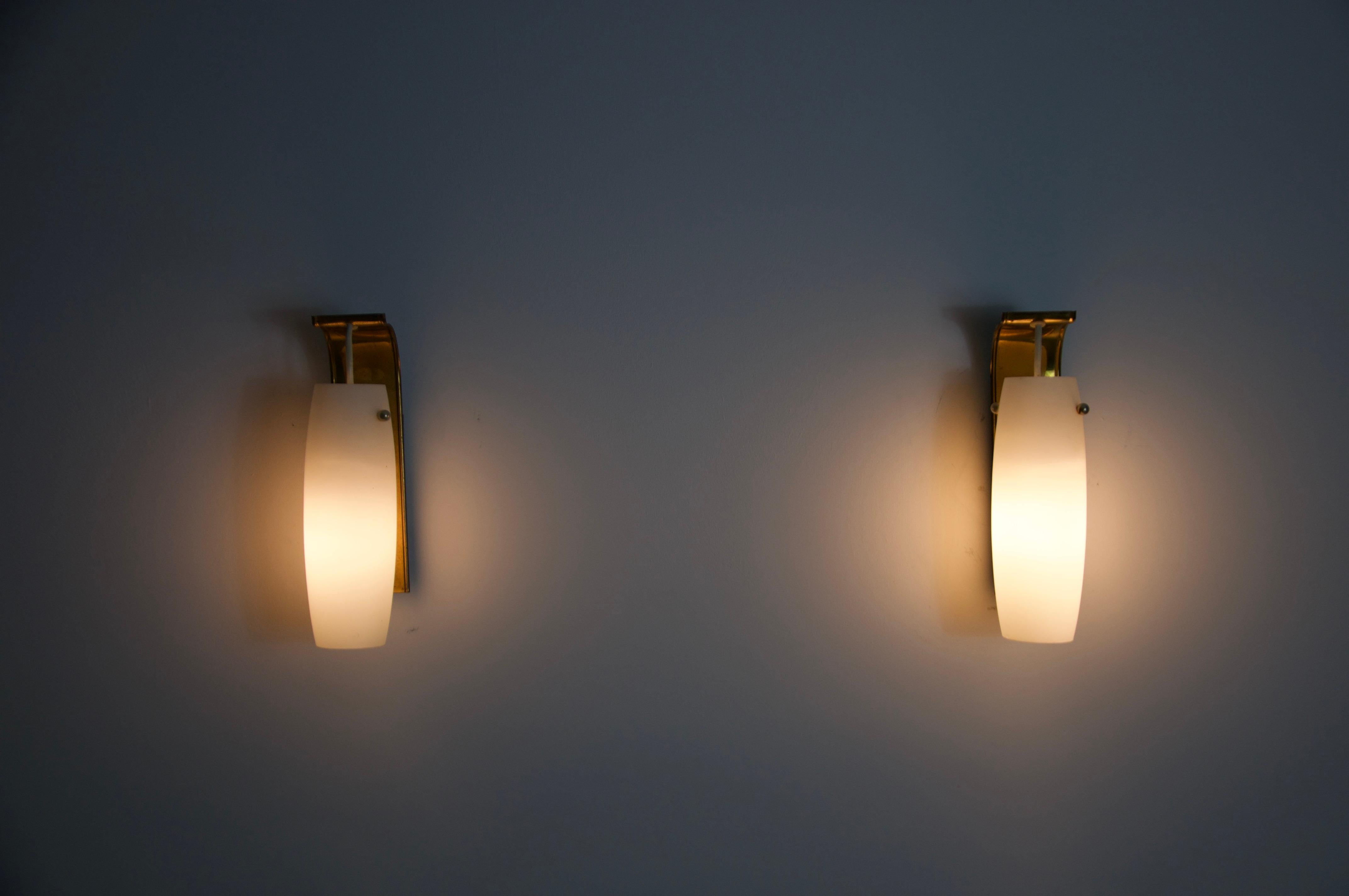 These elegant Limburg wall lamps from the 1960s combine high-quality design with precise German craftsmanship. Each lamp features a curved, rectangular brass body that is mounted to the wall. A sleek, cylindrical opal glass shade is suspended from a