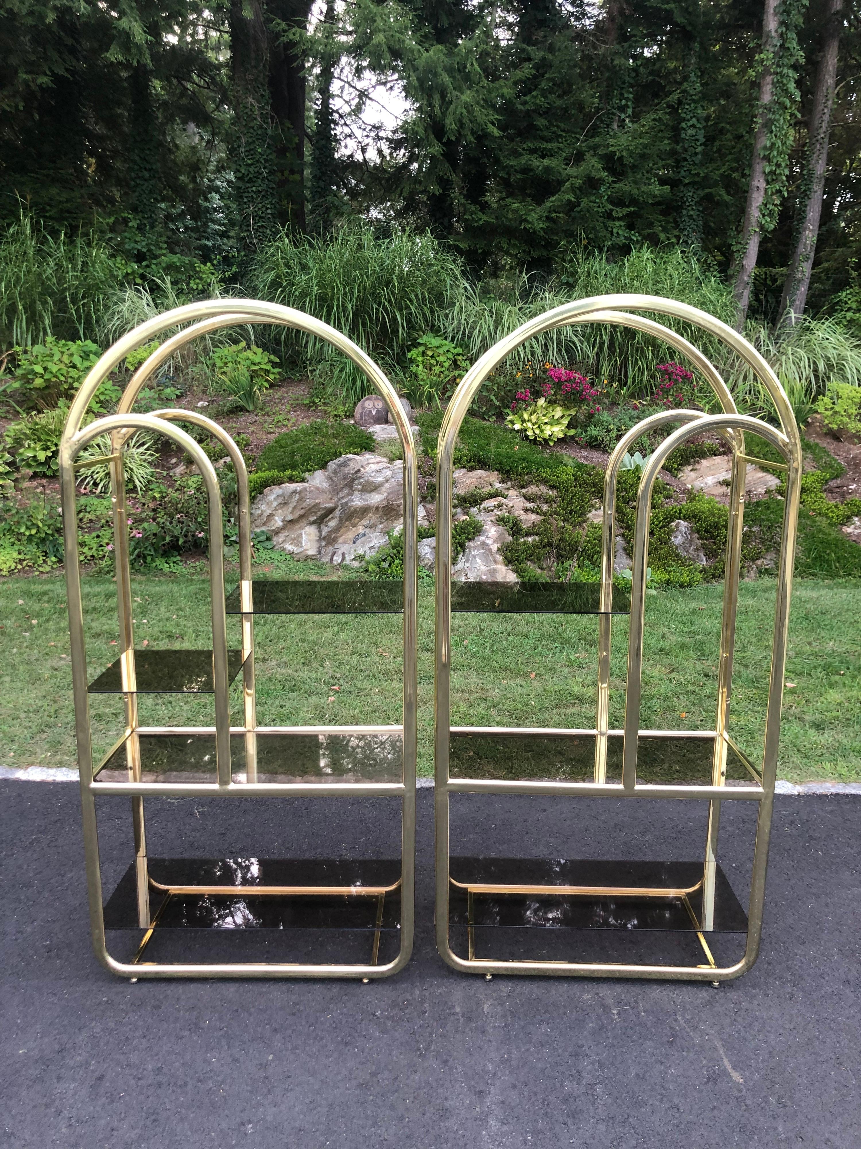 Pair of brass and smoked glass etageres. Post Modern 1980's Hollywood Regency style. Missing one smoked glass shelf in photos that has since been replaced. More photos available. Art deco curved shapes make up this beauty.