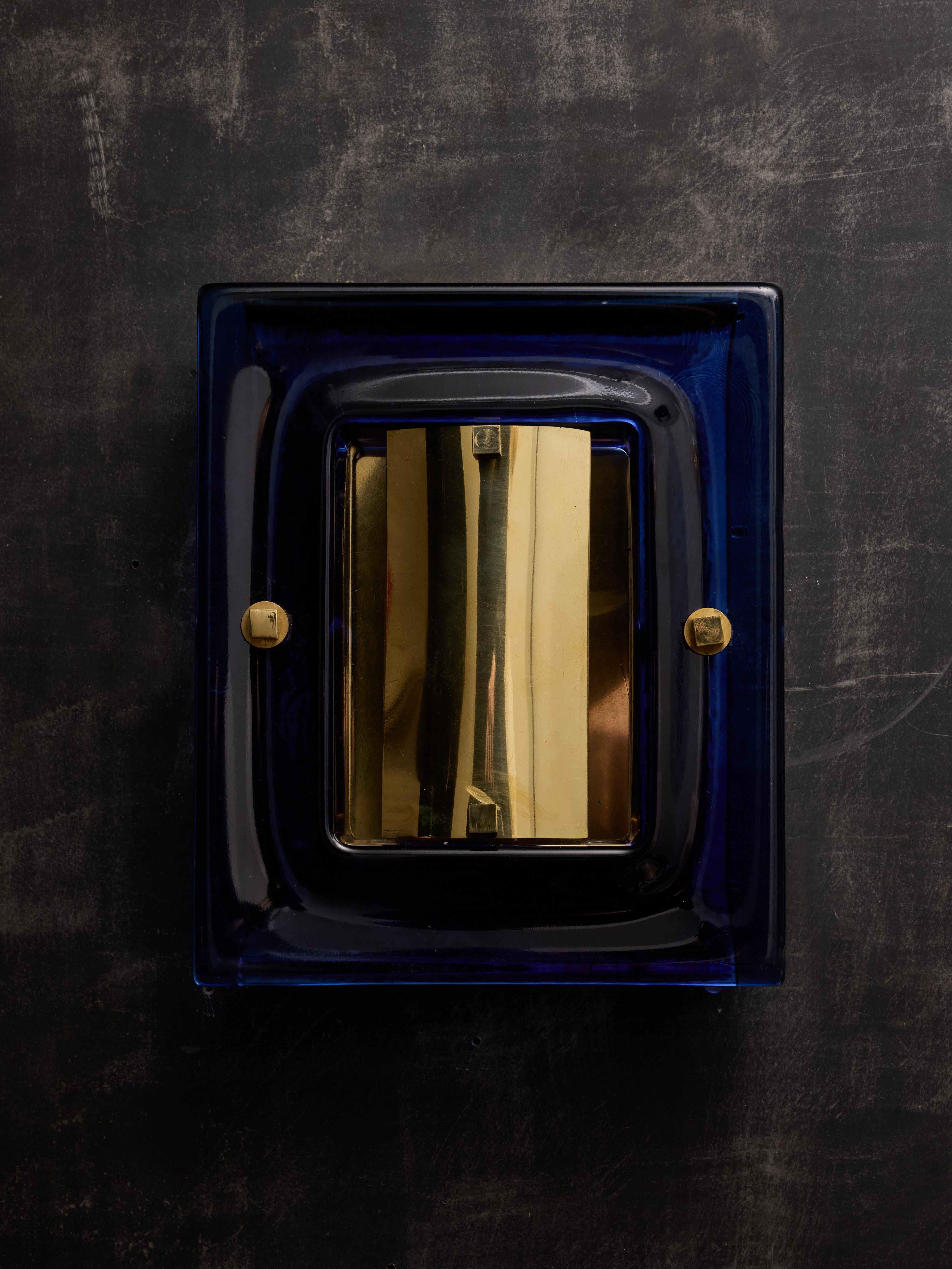 A pair of square wall sconces featuring a brass structure and a thick glass frame in vivid blue. At the center, a single light source is elegantly concealed by a curved brass sheet, allowing the illumination to shine through the glass.
Two pairs