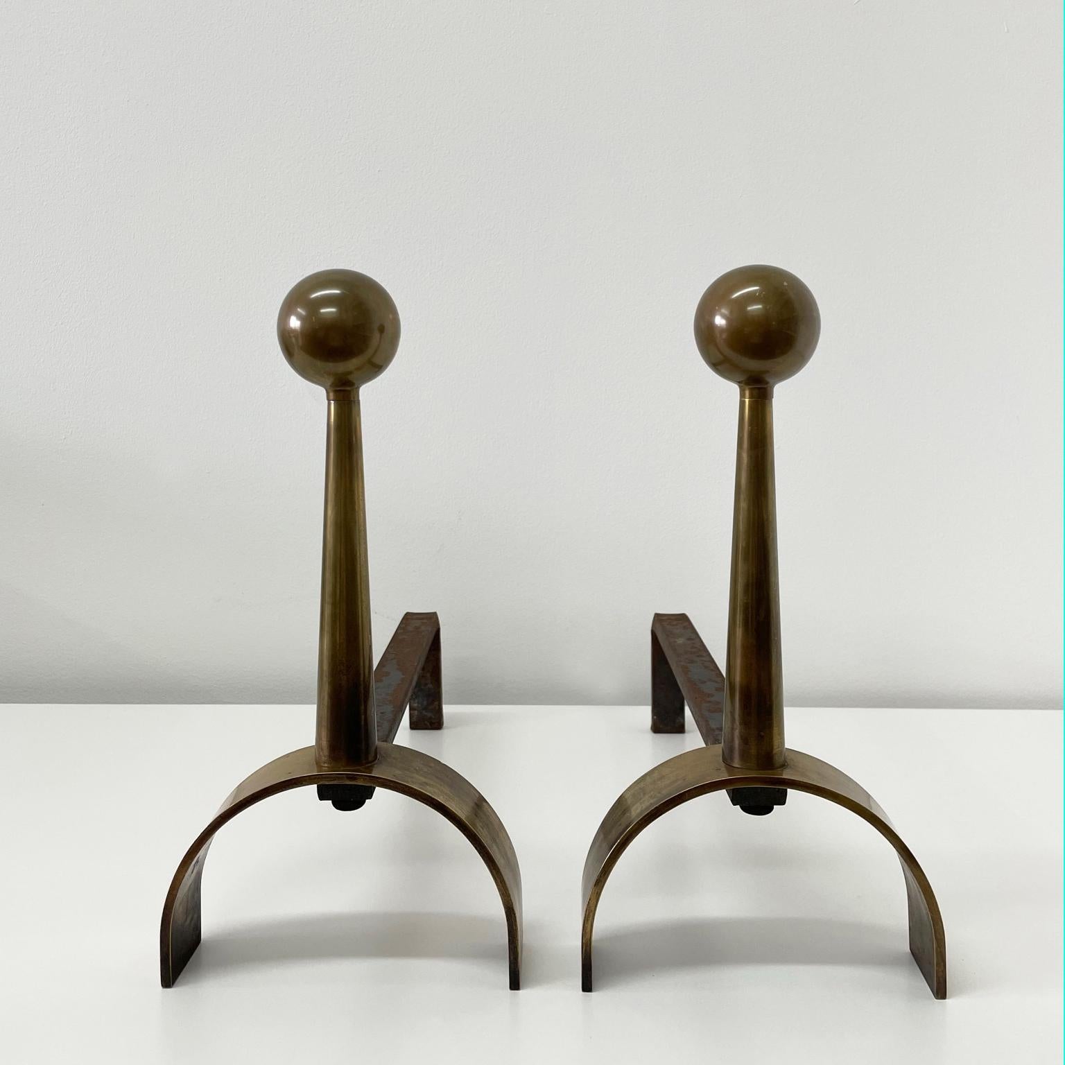 These weighty brass andirons are beautifully proportioned and show a warm patina. A graceful arch provides the base for a nicely tapered stem which terminates in a ball finial. Their pared down form is clean and unobtrusive yet with a presence all