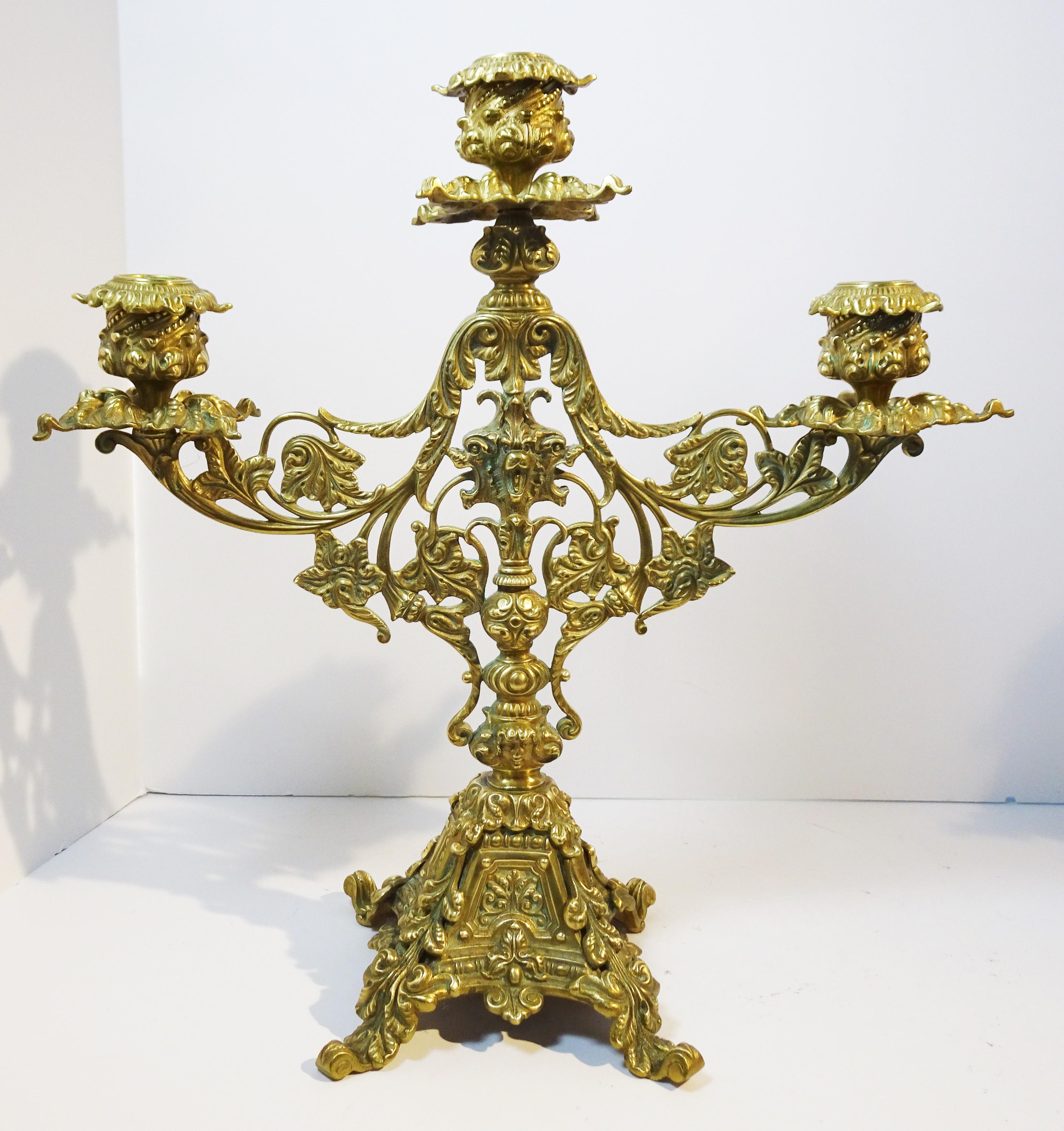 This Baroque-style pair of candelabra are elaborately decorated with leafy scrolls, stems and blossoms on all the surfaces, and are centered by a three. -headed lion over a ball-segmented shaft, the base of which is anchored by a human head on