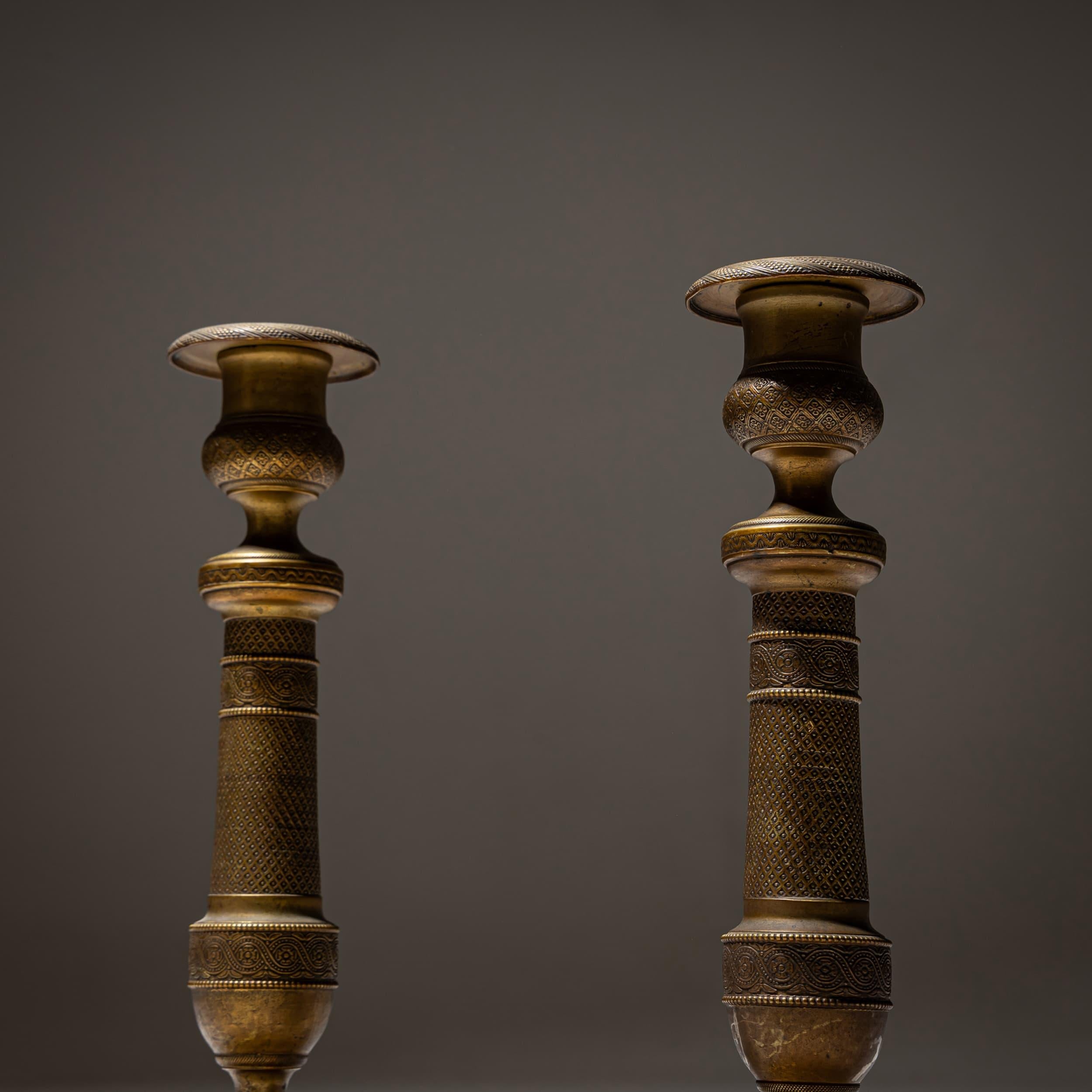 Coppia di candelieri in ottone con basi rotonde, fusti a balaustro e prese cilindriche. La superficie è finemente cesellata con motivi geometrici e bande ornamentali circolari. 