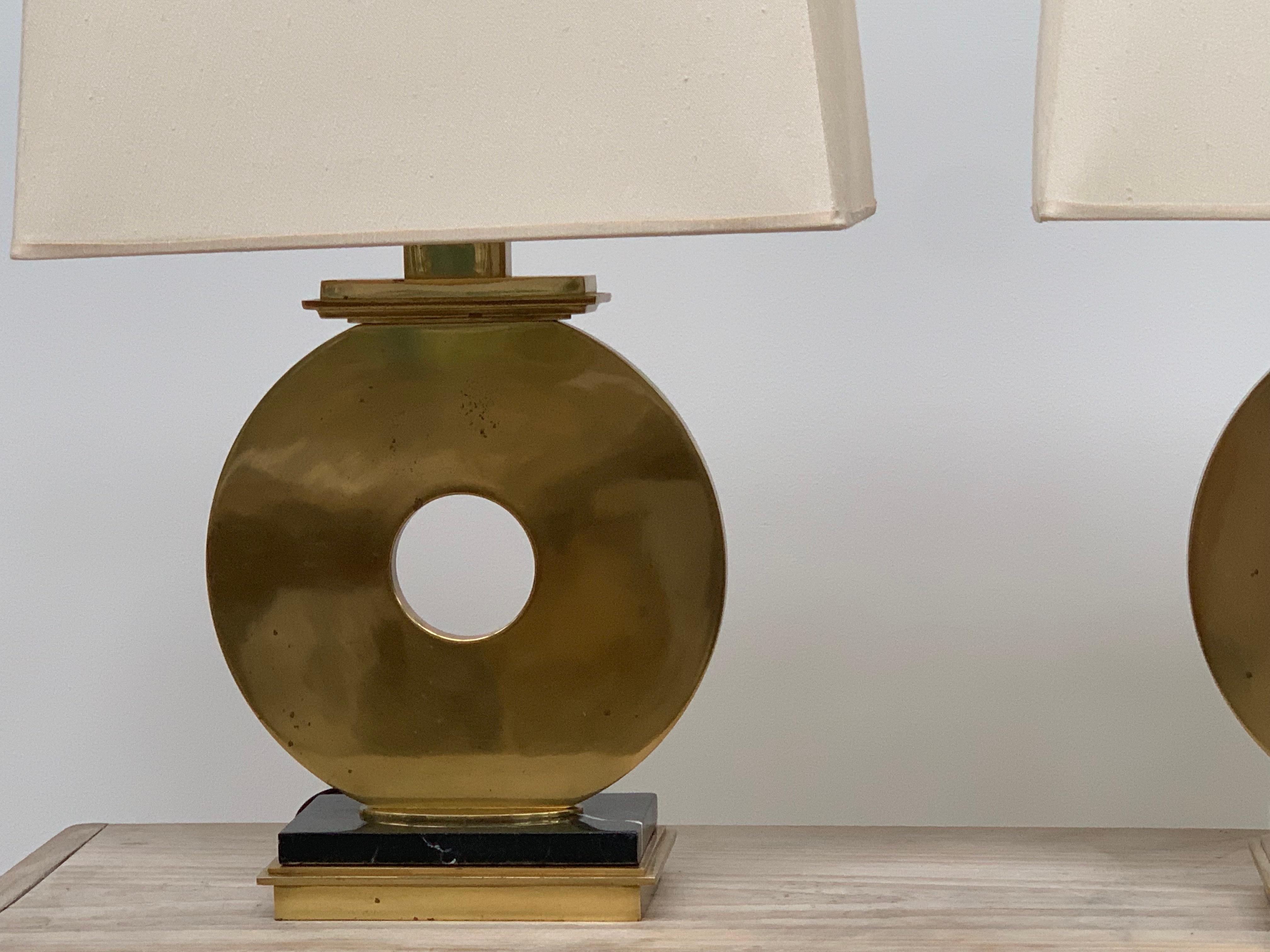 Pair of heavy brass lamps by Robert Abbey. Narrow in profile, round design of an imposing circle of brass on a black marble base. With original shades, signature on harps.