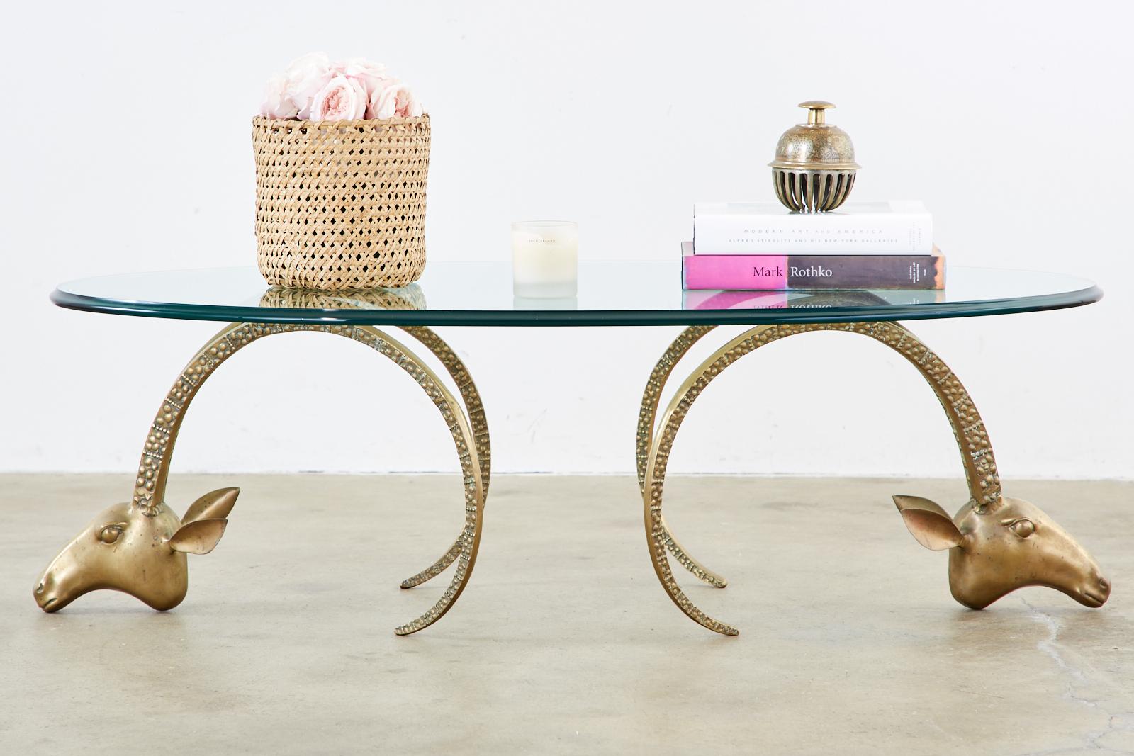 Iconic pair of brass rams heads or Ibex made in the manner and the style of Alain Cheruet. Features a large oval pane of thick, beveled glass. The coffee or cocktail table from the Hollywood Regency era has a lovely aged brass patina on the finish.