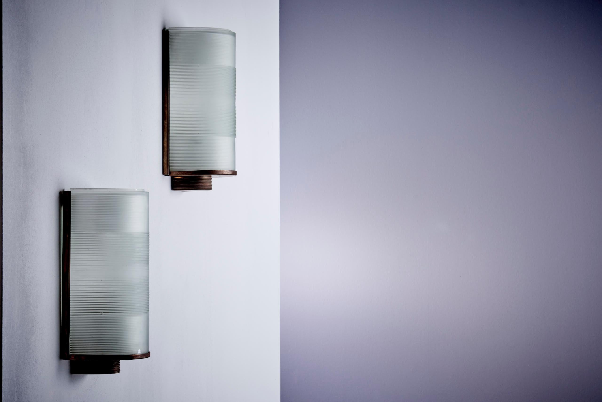 Set of two vintage wall sconces with frosted ribbed glass diffusers and patinated brass frames. The gently curved design and grooved detailing give these lights a sleek, Art Deco-inspired look. They emit a warm, diffused glow and work beautifully in