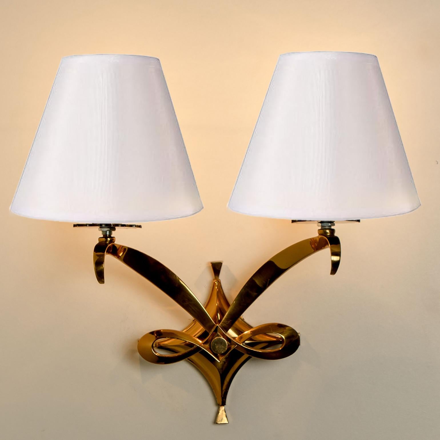 Regal brass Leleu wall sconces. Each light fixture consists of curved gold rods, with a white lampshade on top. Mounted on a gold-plated brass frame. The rods refract light beautifully. Multiple options are available in terms of lampshades, please