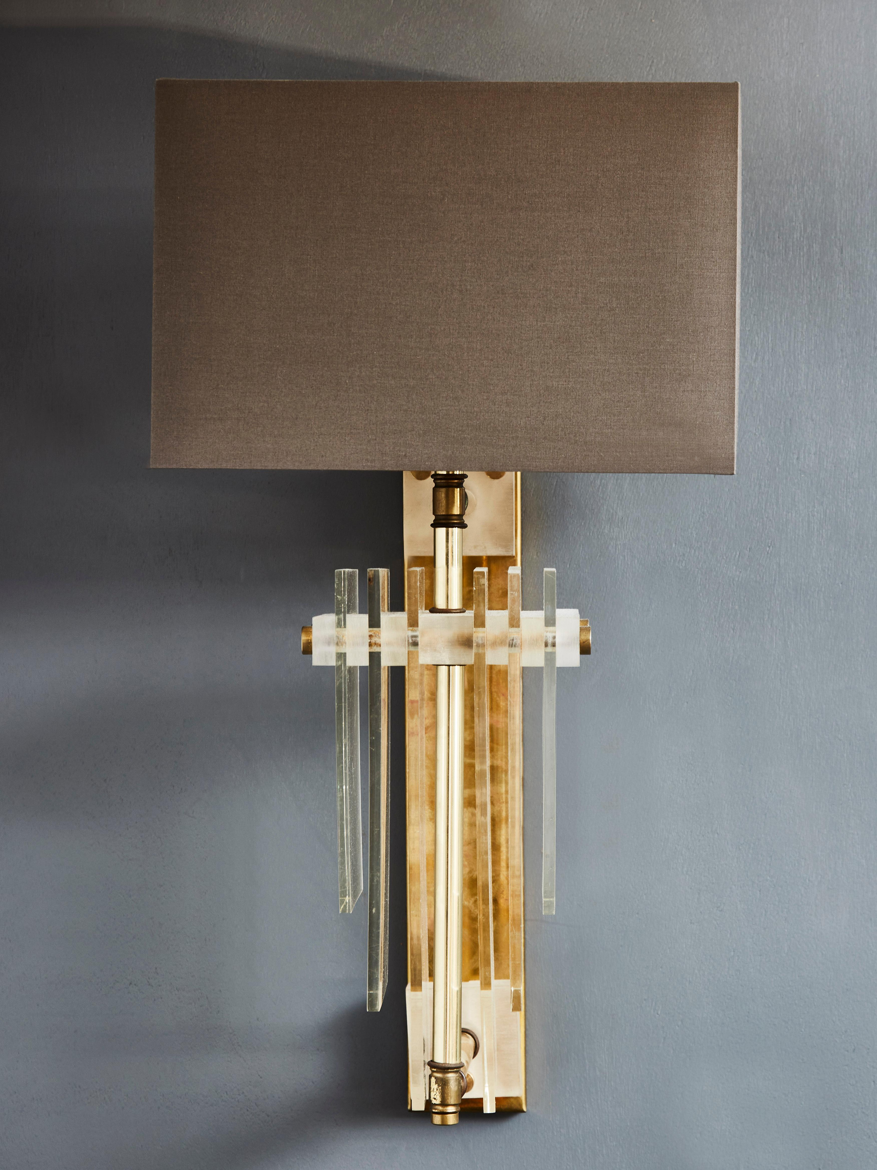 Pair of wall sconces made of brass with decorative plexiglass slabs toped with rectangular grey shades.