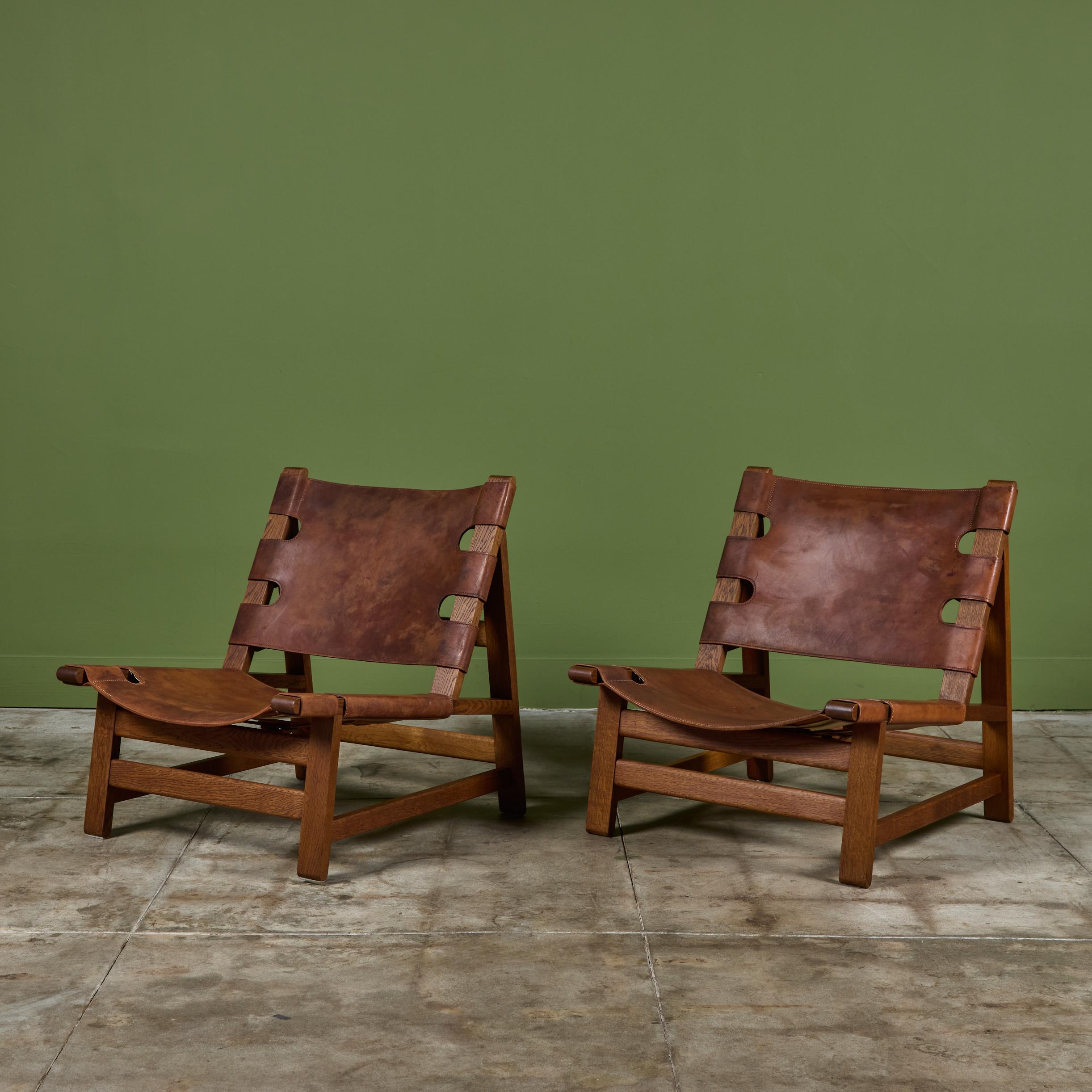 Pair of oak and leather lounge chairs by Børge Mogensen for Fredericia Furniture c.1960s, Denmark. The chairs have a low wide leather sling seat in cognac and the seat backs showcase brass buckle details along the back and at the bottom of the