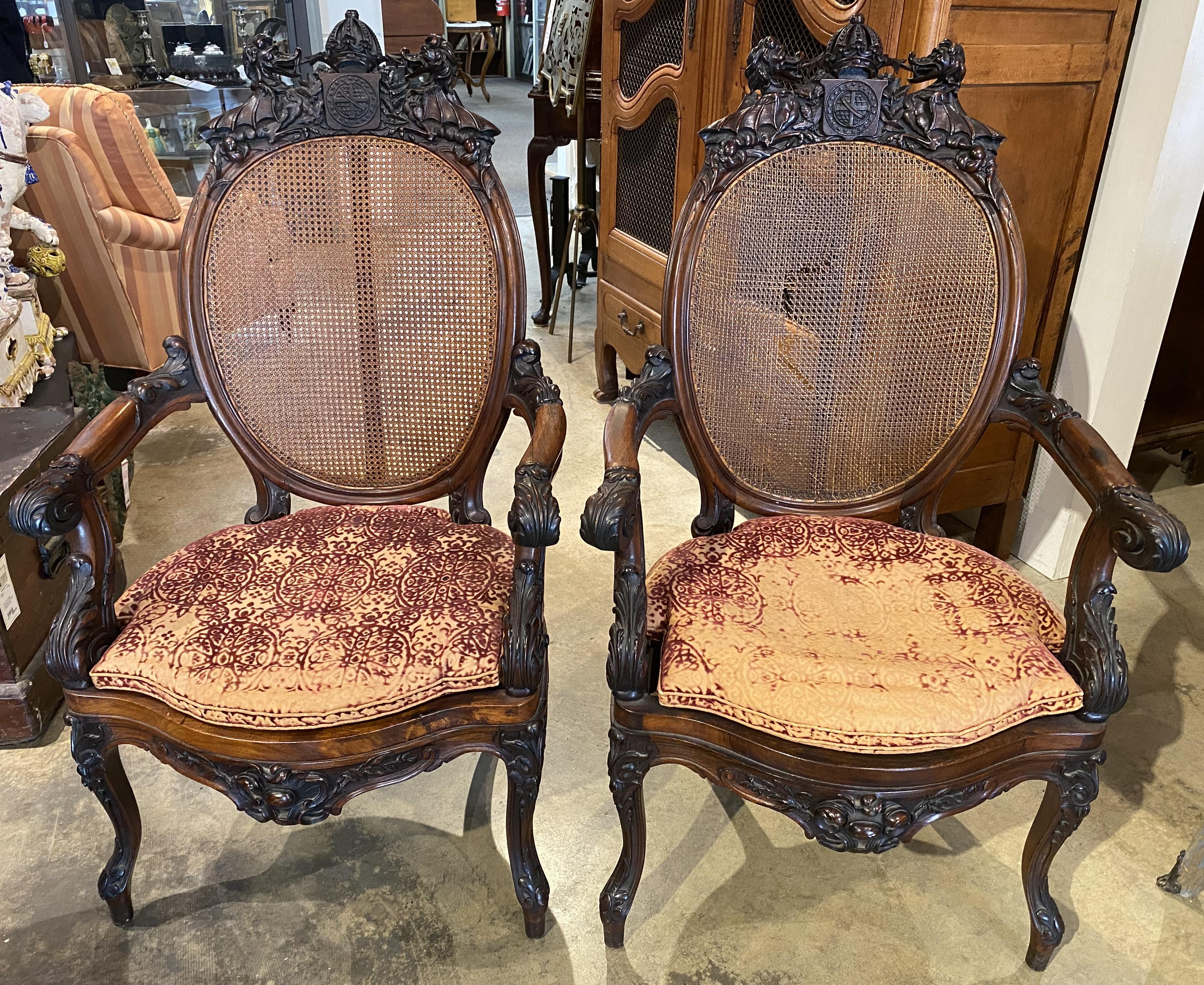 A fine pair of British walnut open armchairs with detailed carved crown & dragon crests surmounting foliate carved arms, seats and legs accented with caned backs and seats, with thin upholstered custom cushions. The pair date to the 19th century and