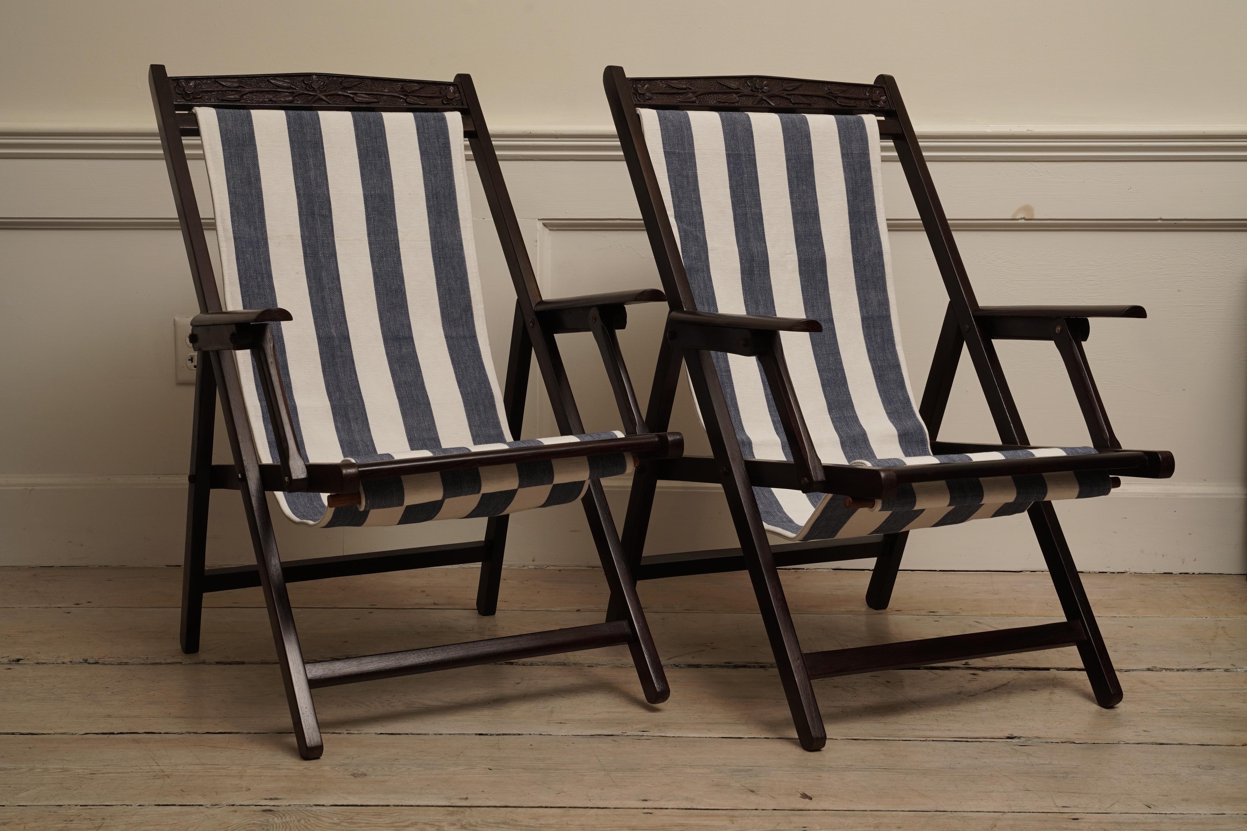 A pair of teak, British Campaign folding chairs with a striped canvas sling seat with arm rests. The fabric has been replaced. Carved vine motif at the crest. Mid 1900's. Canvas can be easily swapped out for cleaning or replacing. Great for