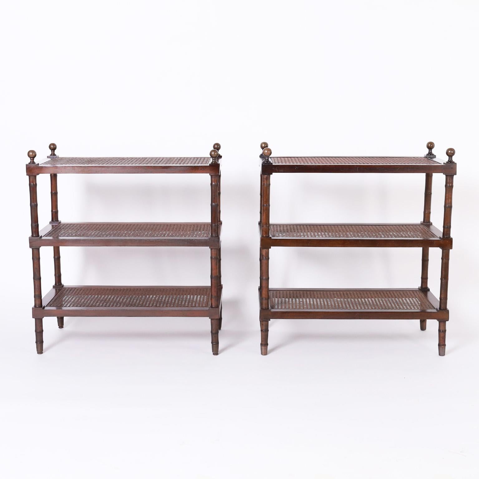 Standout pair of British colonial style stands or tables crafted in mahogany having brass finials, three caned tiers with the top tier over glass, and faux bamboo supports and legs.