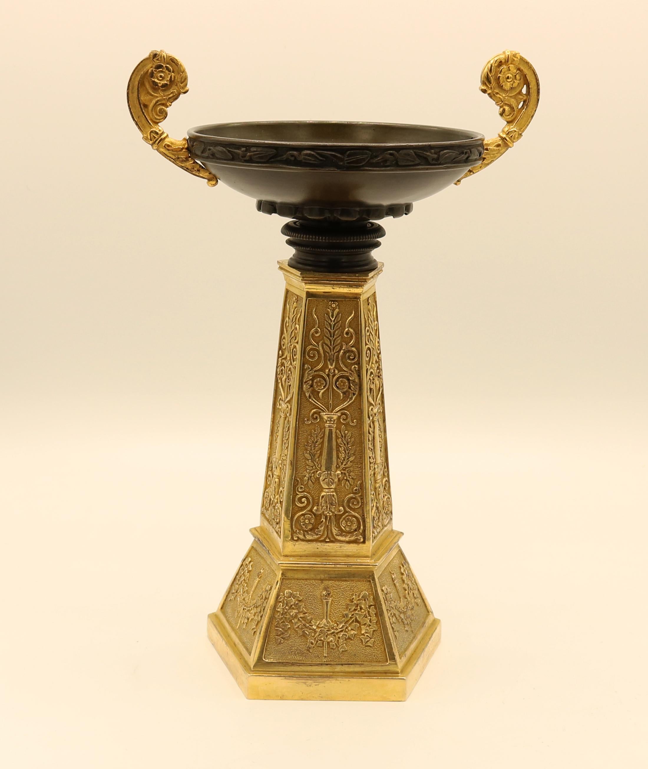A pair of mid-19th century bronze and ormolu Tazzas of unusual design, the bowls having leaf and berry decorated edges with scroll handles above hexagonal panelled columns decorated with stylised candelabra supported on plinth bases with leaf