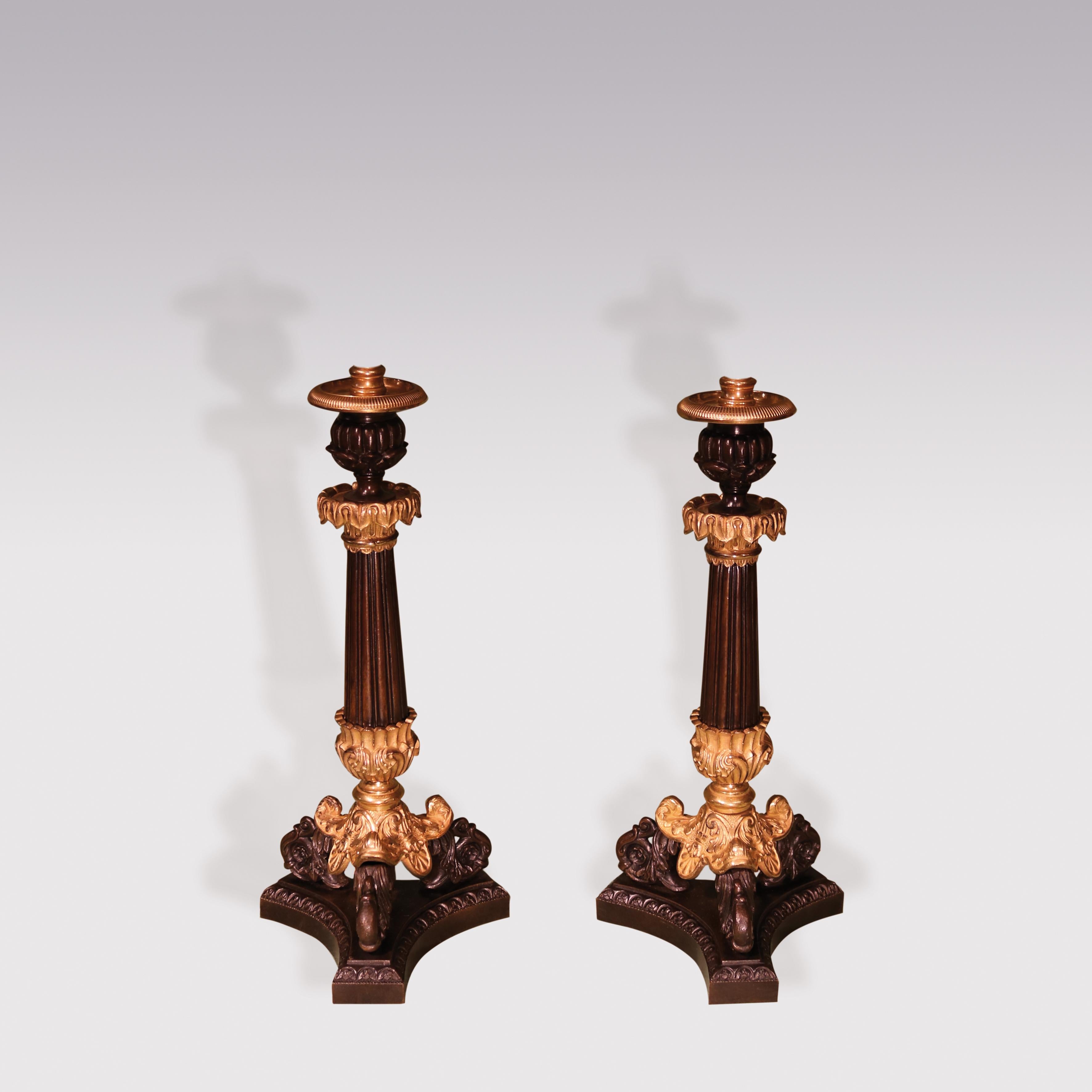 A pair of early 19th century bronze and Ormolu candlesticks, having leaf-decorated nozzles above reeded, tapering stems raised on acanthus scrolled triform feet ending on concave platform base. (Now converted to Lamps).
