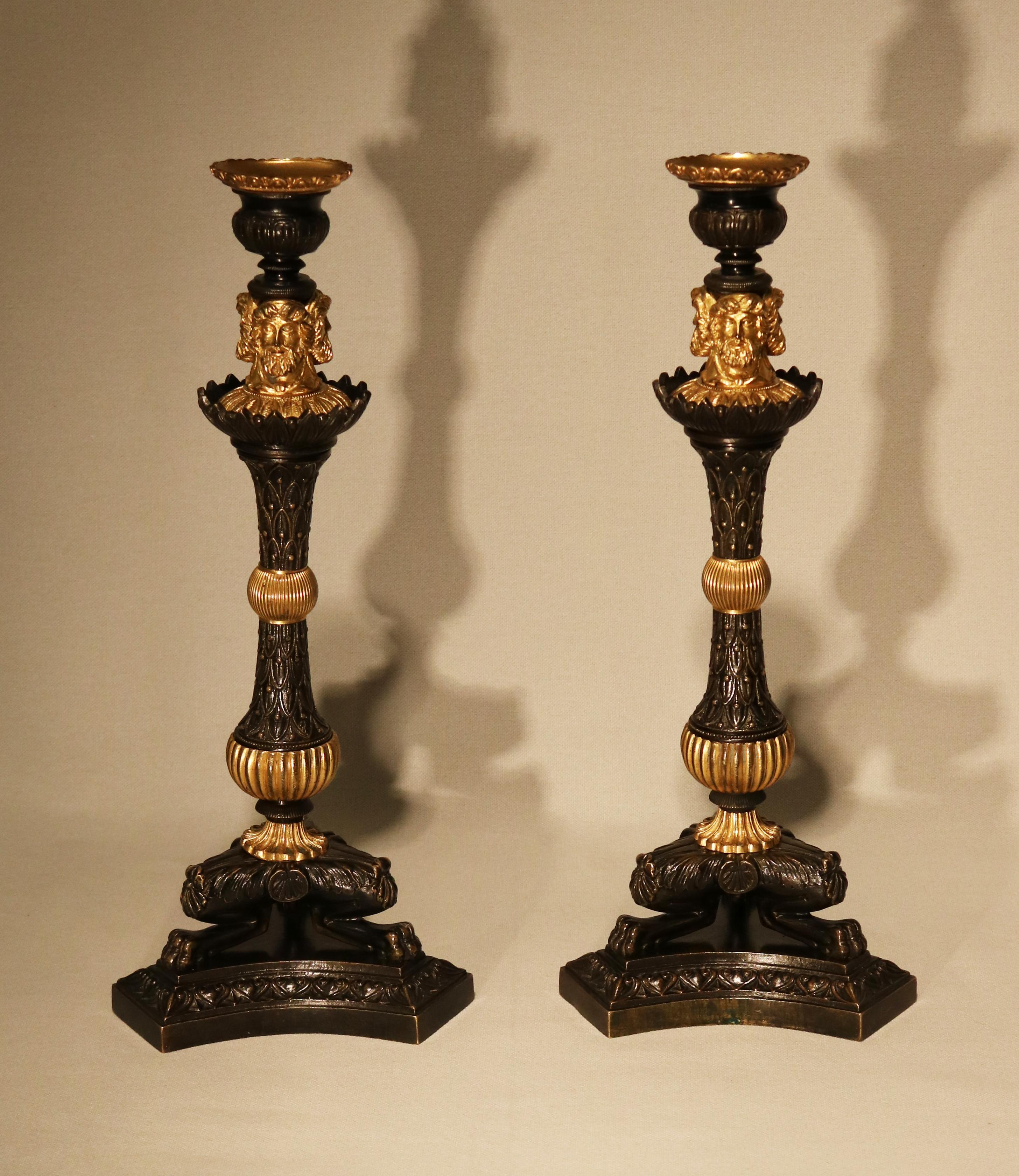A pair of early 19th century bronze and ormolu candlesticks having urn-shaped sconces above triple above triple classical heads raised on leaf and berry tapering stems centred with reeded ball sections, raised on triform legs with lion’s paw feet