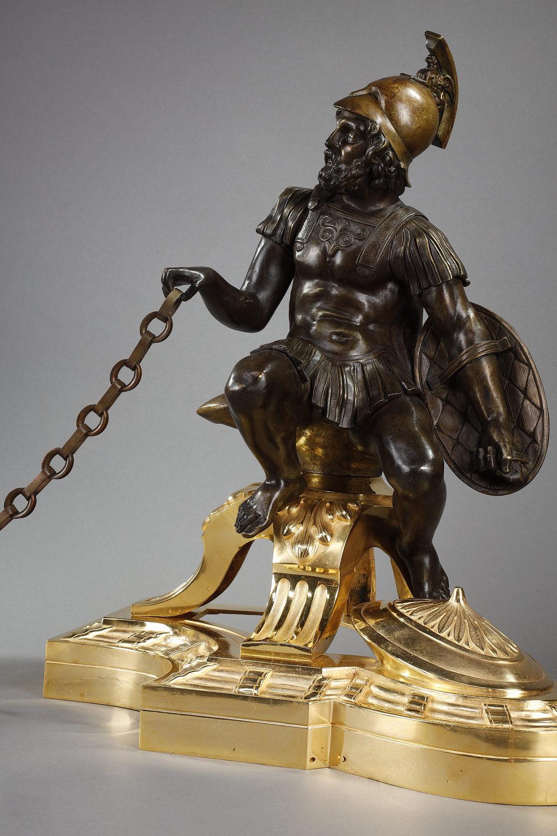 Une paire de chenets en bronze patiné et doré représentant un soldat et un mendiant. L'une d'elles montre un soldat portant un casque macédonien avec un cimier en forme de sphinx, une cuirasse moulant sa forme et tenant un bouclier. L'autre montre