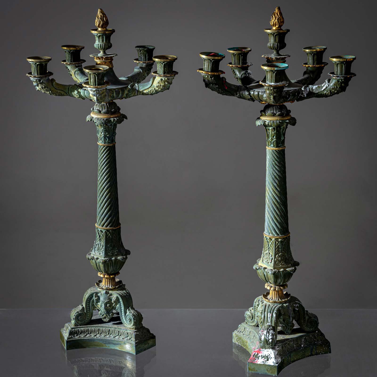 Coppia di candelieri in bronzo patinato verde con cinque bracci e un'urna centrale con motivo a fiamma. Il fusto contorto conduce alla base a trifoglio con motivi a rocaille e foglie.