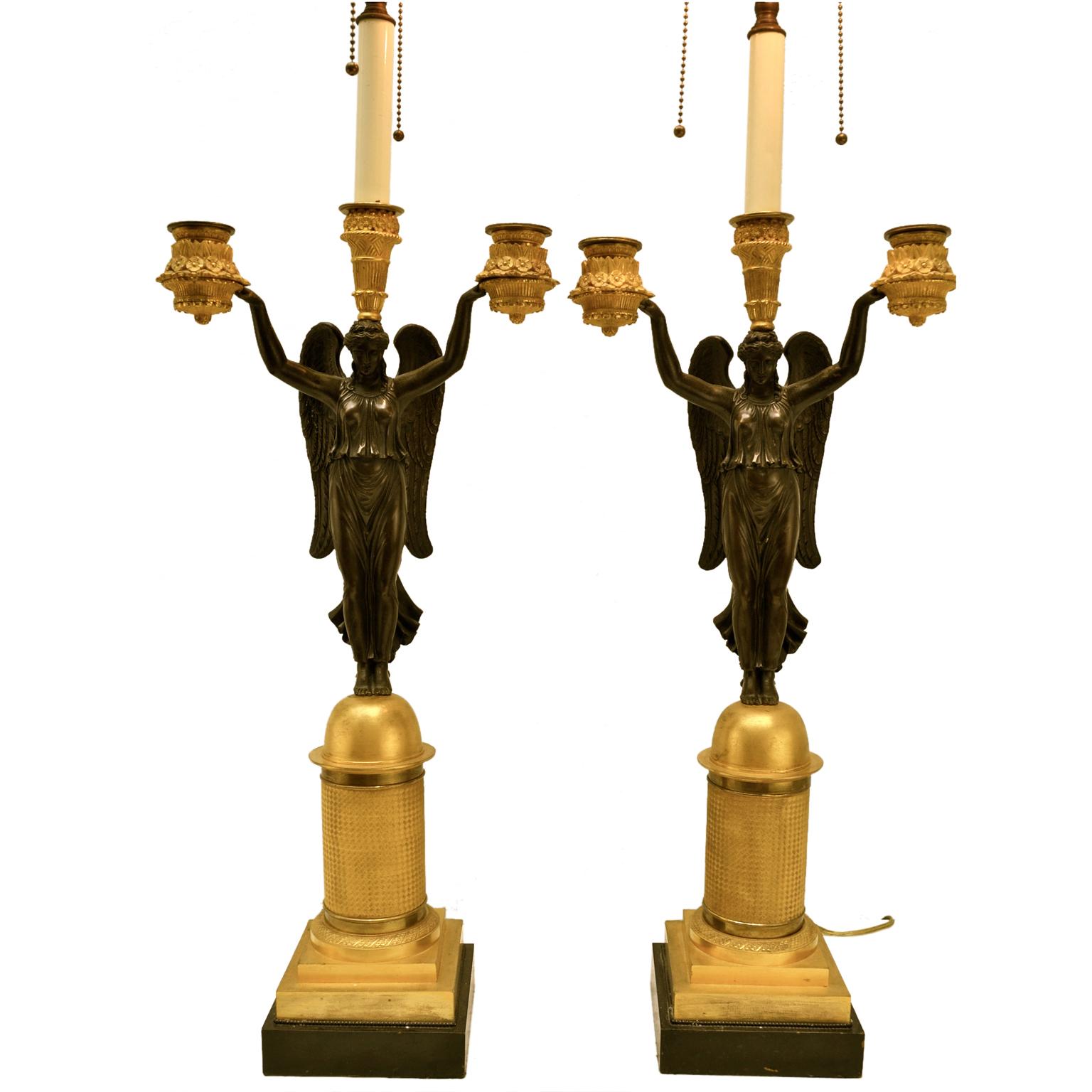 A period pair of French Empire candlestick or lamps, showing a patinated bronze winged Victory her arms aloft holding two decorative gilded candle nozzles resembling cornucopia, her head supports a third candle nozzle. She stands on a gilded half