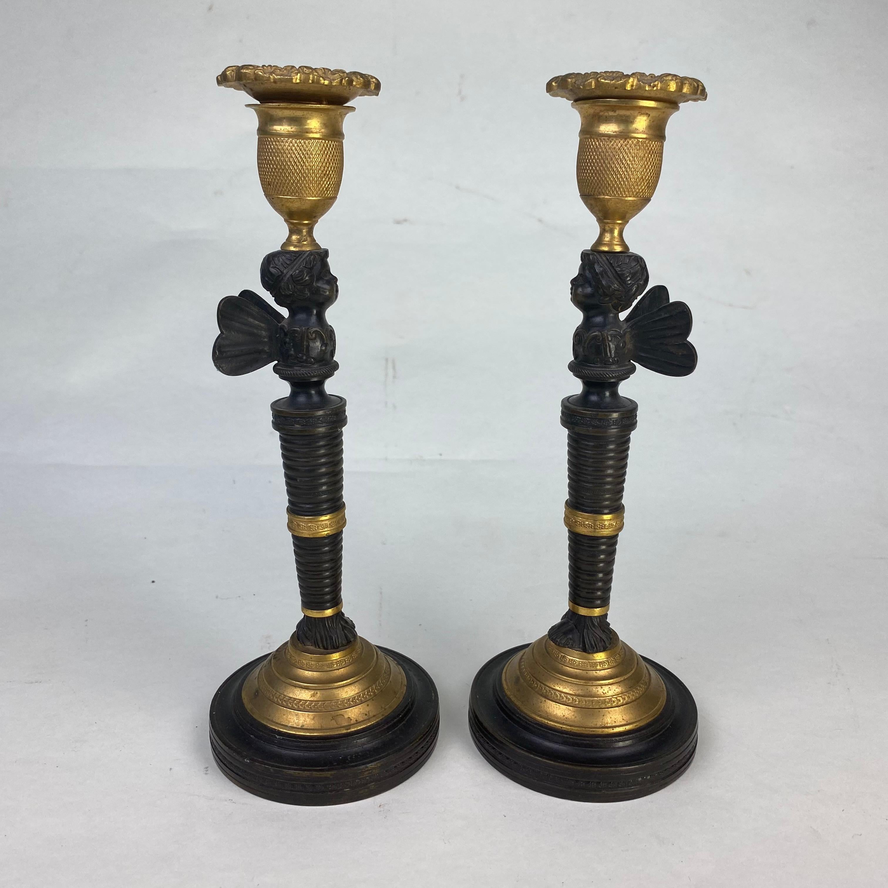 A good quality pair of bronze and ormolu candlesticks with ormolu socles on decorated brionze bases supporting tapered ringed columns with bronze winged putti and ormolu sconces with removable drip pans.