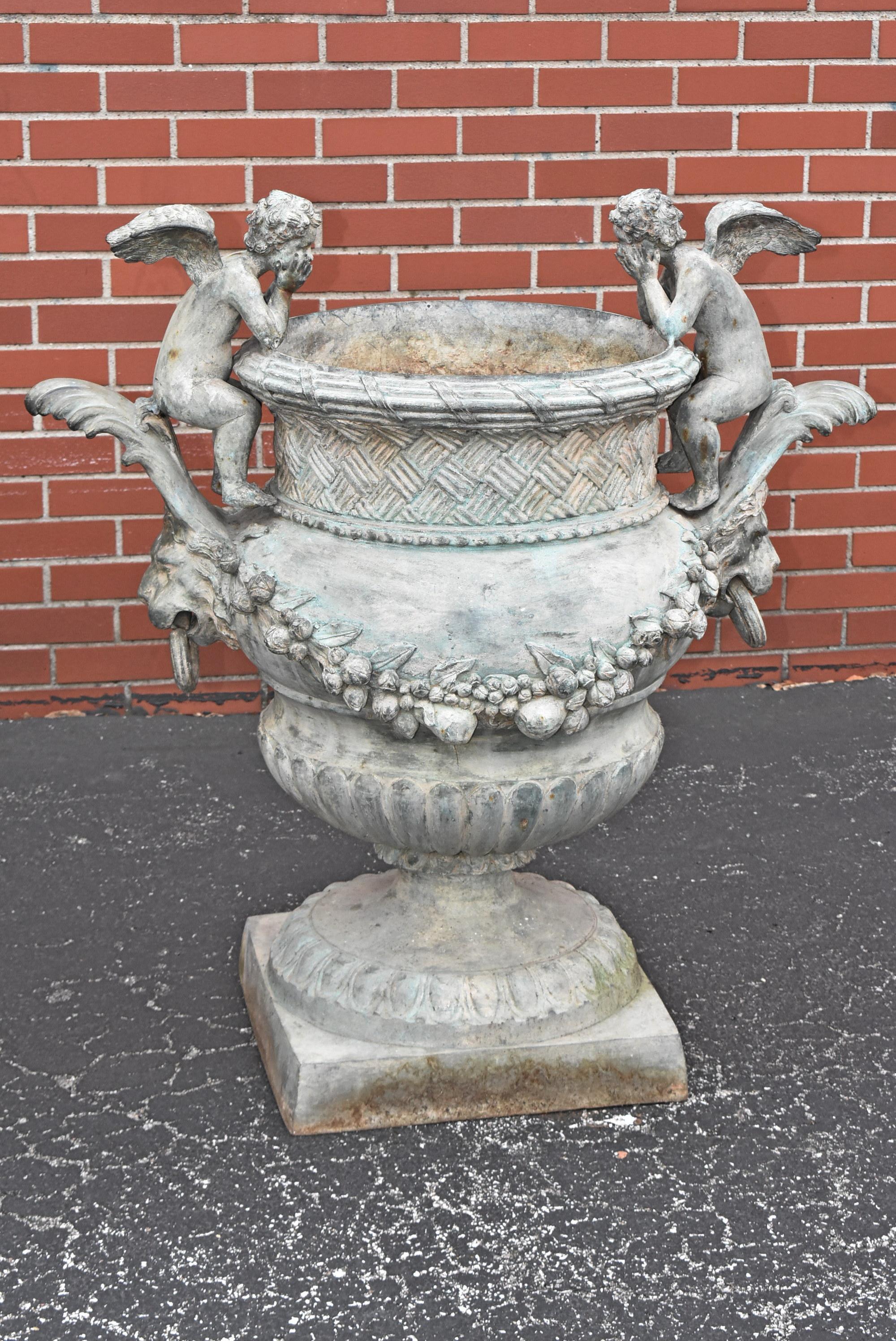 Pair of Bronze Outdoor Urns with Winged Cherubs. Exquisite detail from its basket-woven crown and pondering winged cherub handles to its swags of fruits and flowers. Lion head knockers on each side. Green patina in color on the bronze. Good