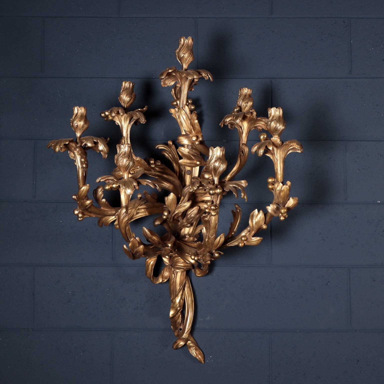 Monumental pair of bronze sconces with seven branches. Two big branches are held by a bow with love knot, they are the sculptural protagonists from which the seven candleholder branches depart. A floral corolla supports a curled chalice shaped as a
