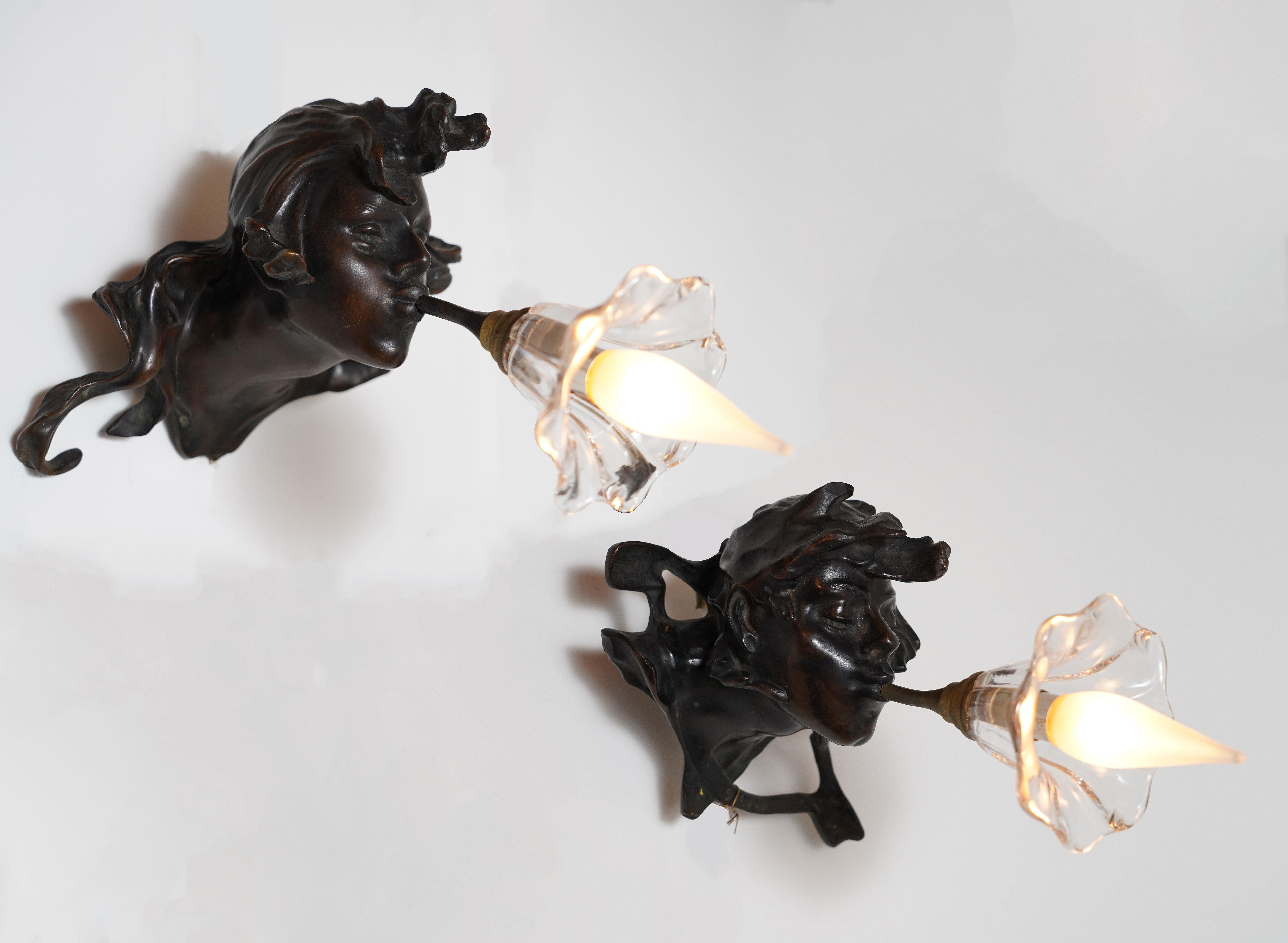 Charmantes Paar patinierter Bronzewandleuchter, die zwei männliche Köpfe darstellen, die in eine leuchtende Glaskrone blasen, die aus ihren Mündern entspringt.

Dieses Modell einer Wandleuchte ist das berühmteste Werk des französischen Bildhauers