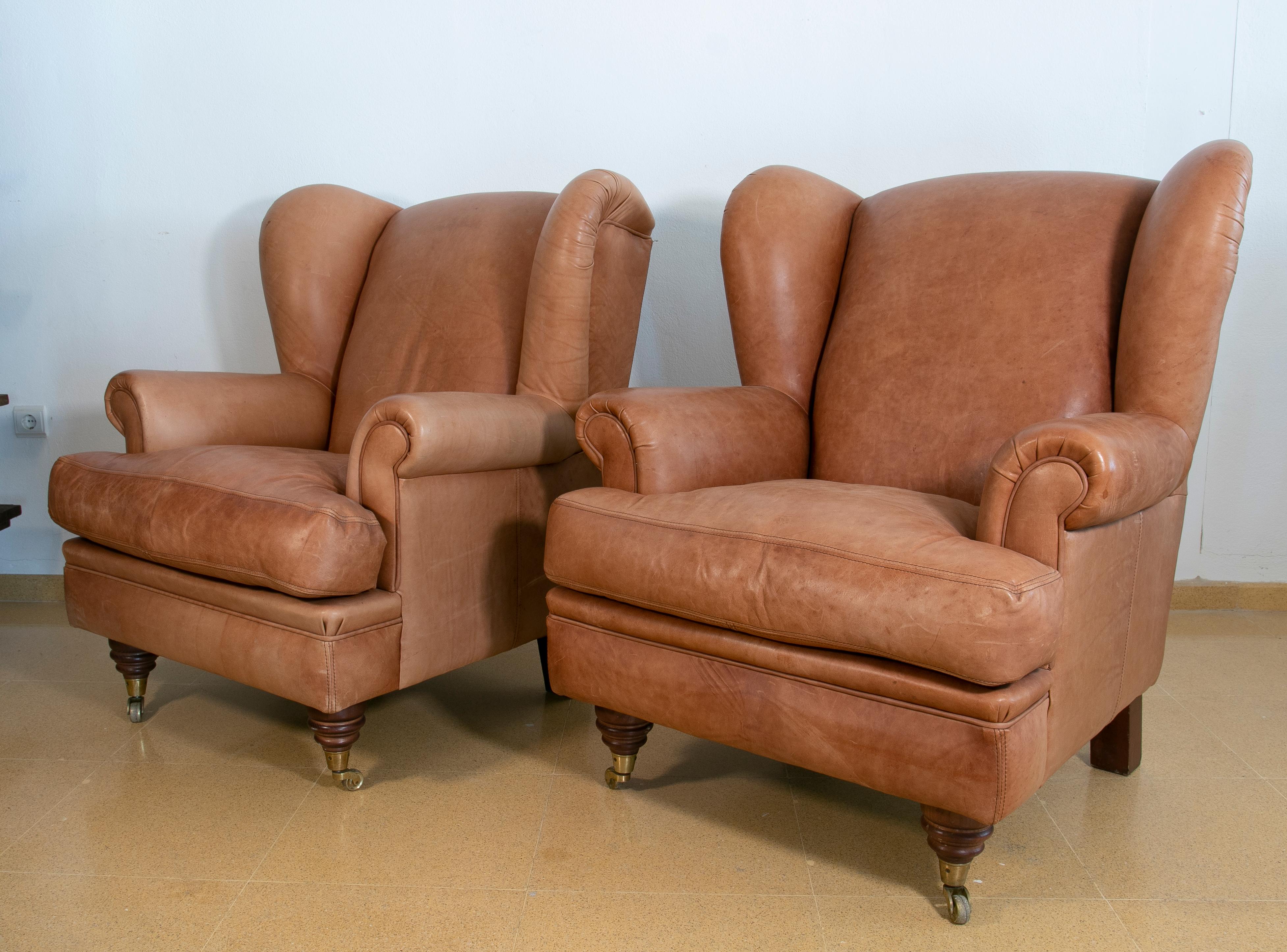 Pair of brown leather armchairs with wooden legs and brass wheels.