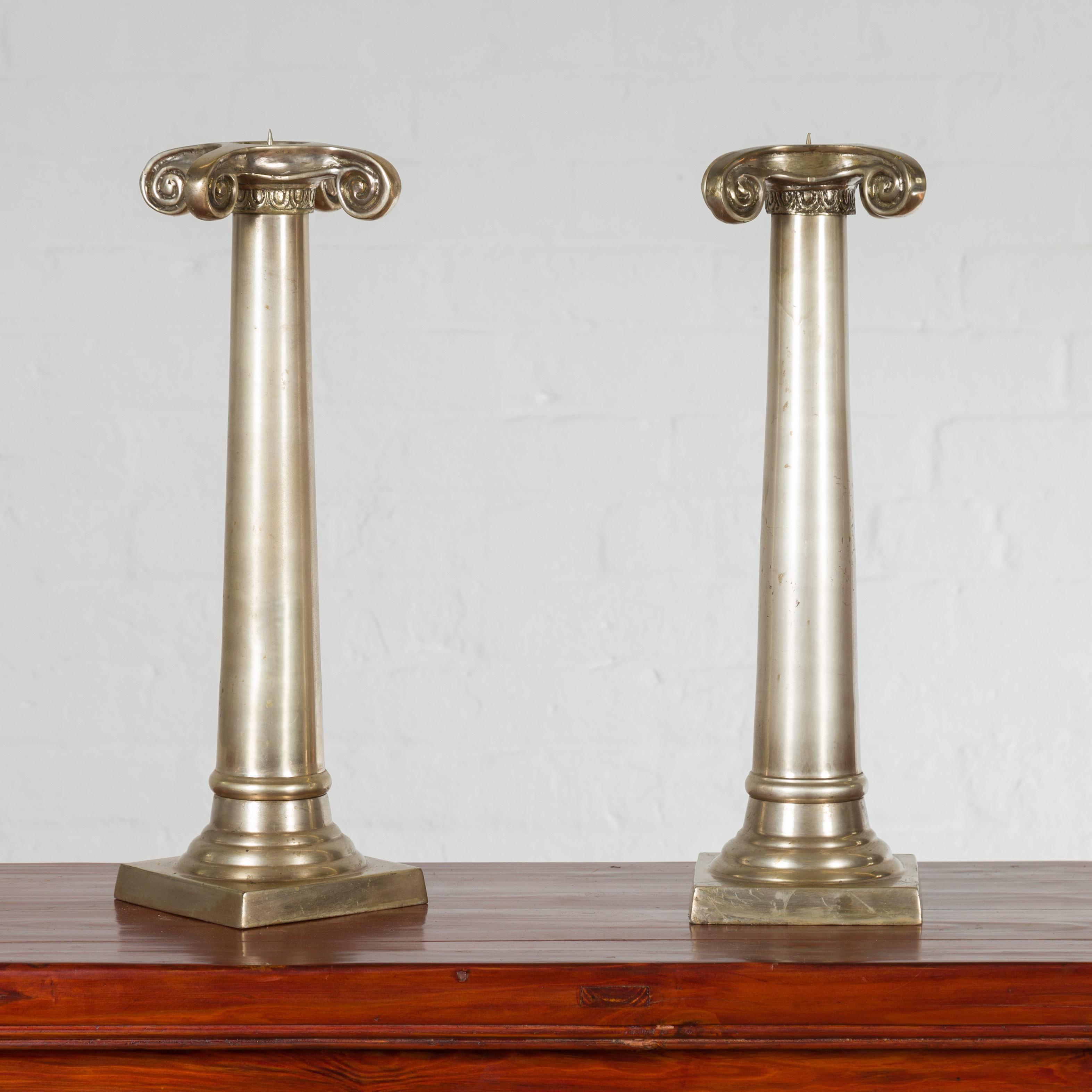 A pair of Thai brushed silver over bronze candlesticks from the mid 20th century, with Ionic capitals. Created during the midcentury period, each of this pair of Thai candlesticks features a sleek column topped with a large Ionic capital. Resting on