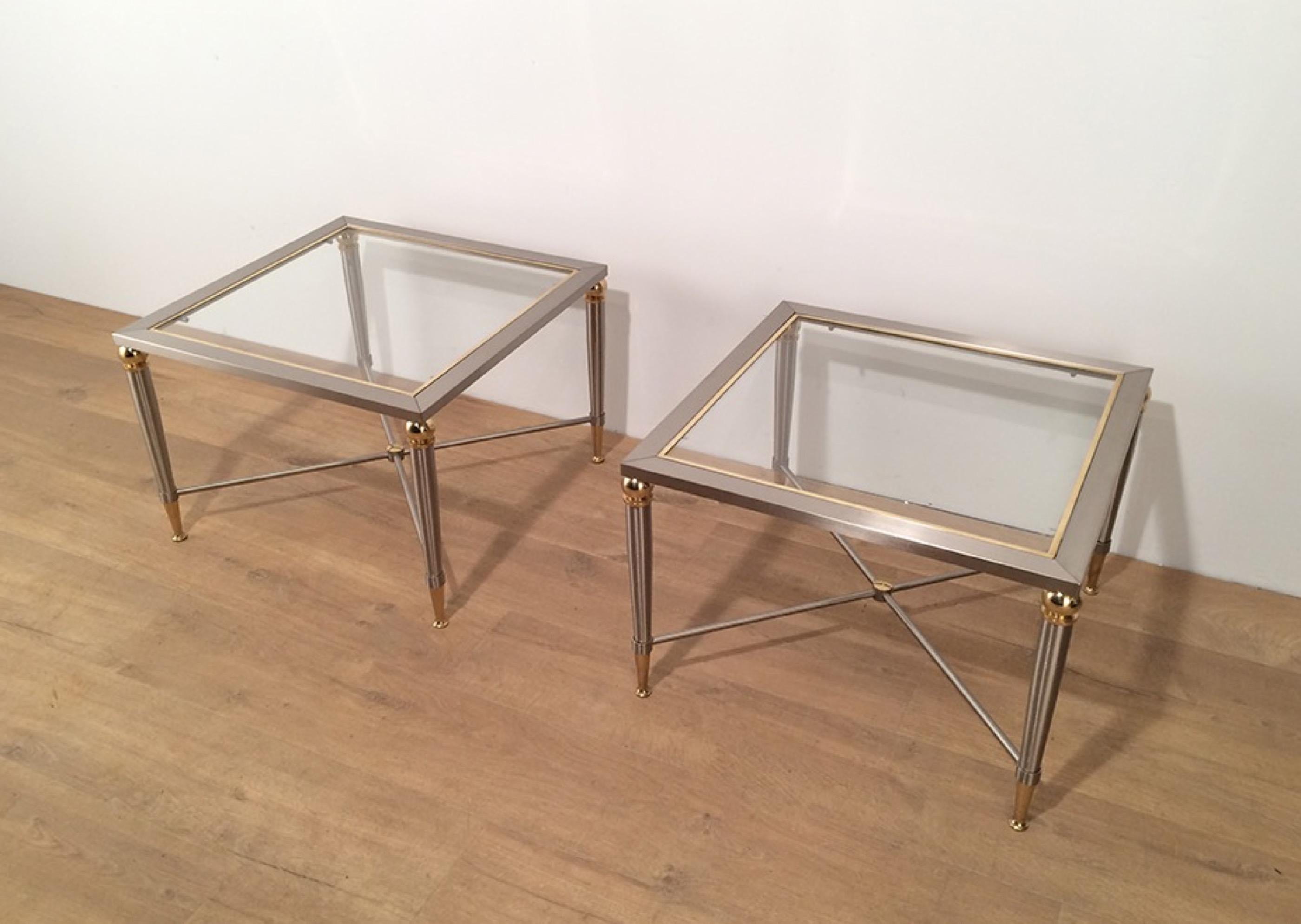 This pair of neoclassical side tables is made of brushed steel and brass. Each end table has glass on top, surrounded by a brass frame and a stretcher on the bottom made of brushed steel cross with a brass decoration on its center. They are very