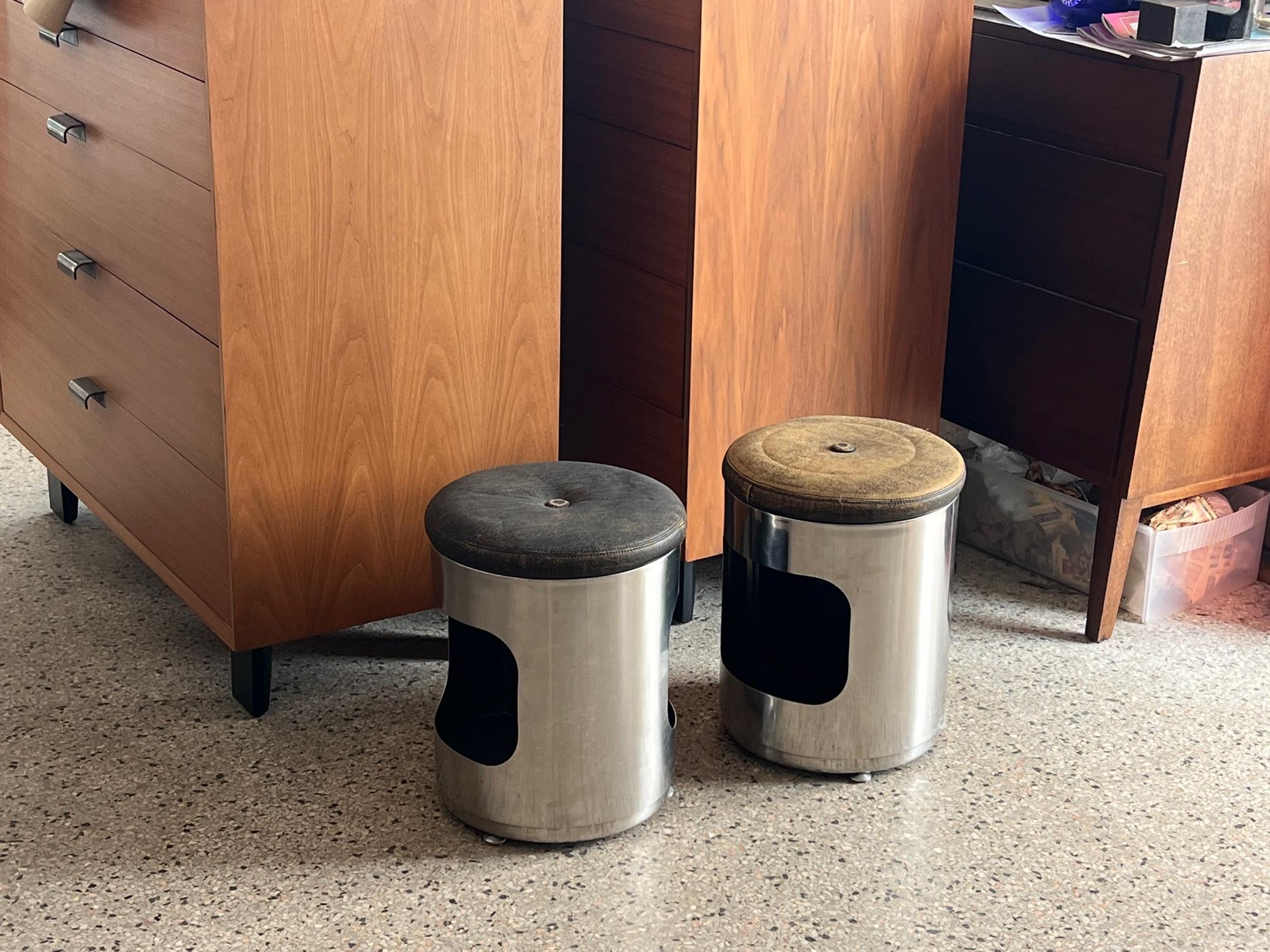 A pair of 1970s very chic, leather/brushed steel upholstered ottomans with symmetric cutouts for storage (magazines, etc. Note the cool conical shaped feet. High quality construction-heavy gauge steel.