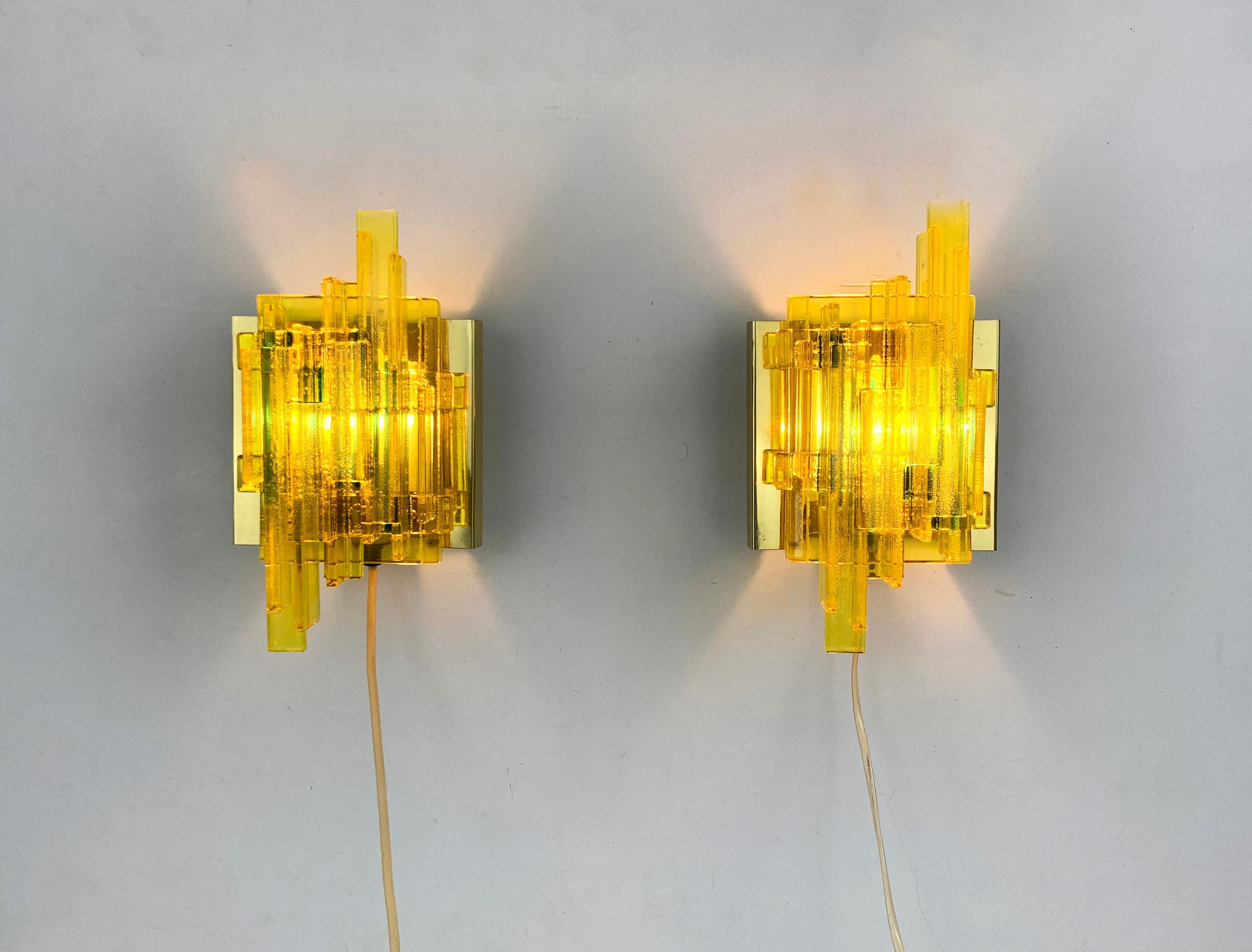 These wall-lamps or sconces were designed by Claus Bolby and manufactured by his own company Cebo Industri in Denmark. 

The lamps come with a composition of yellow acrylics blocs/bars, melted together with inclusion of air bulbs.
When the lamp is