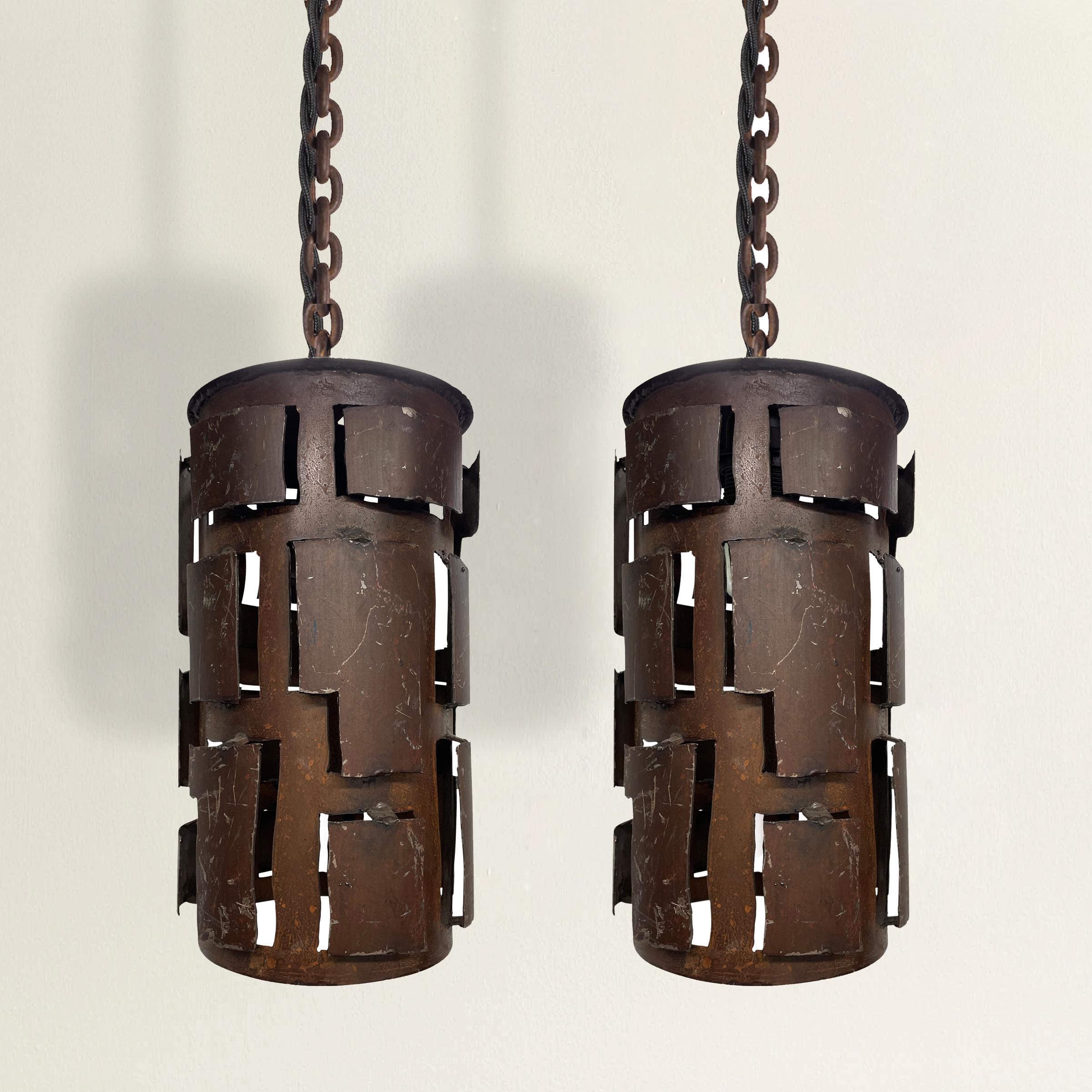 An incredibly chic pair of Brutalist steel pendant lights, each with pierced sides and applied cutouts. Wired for US, with black silk twisted cords. Perfect over a kitchen island, down a hallway, or hung on either side of your bed.