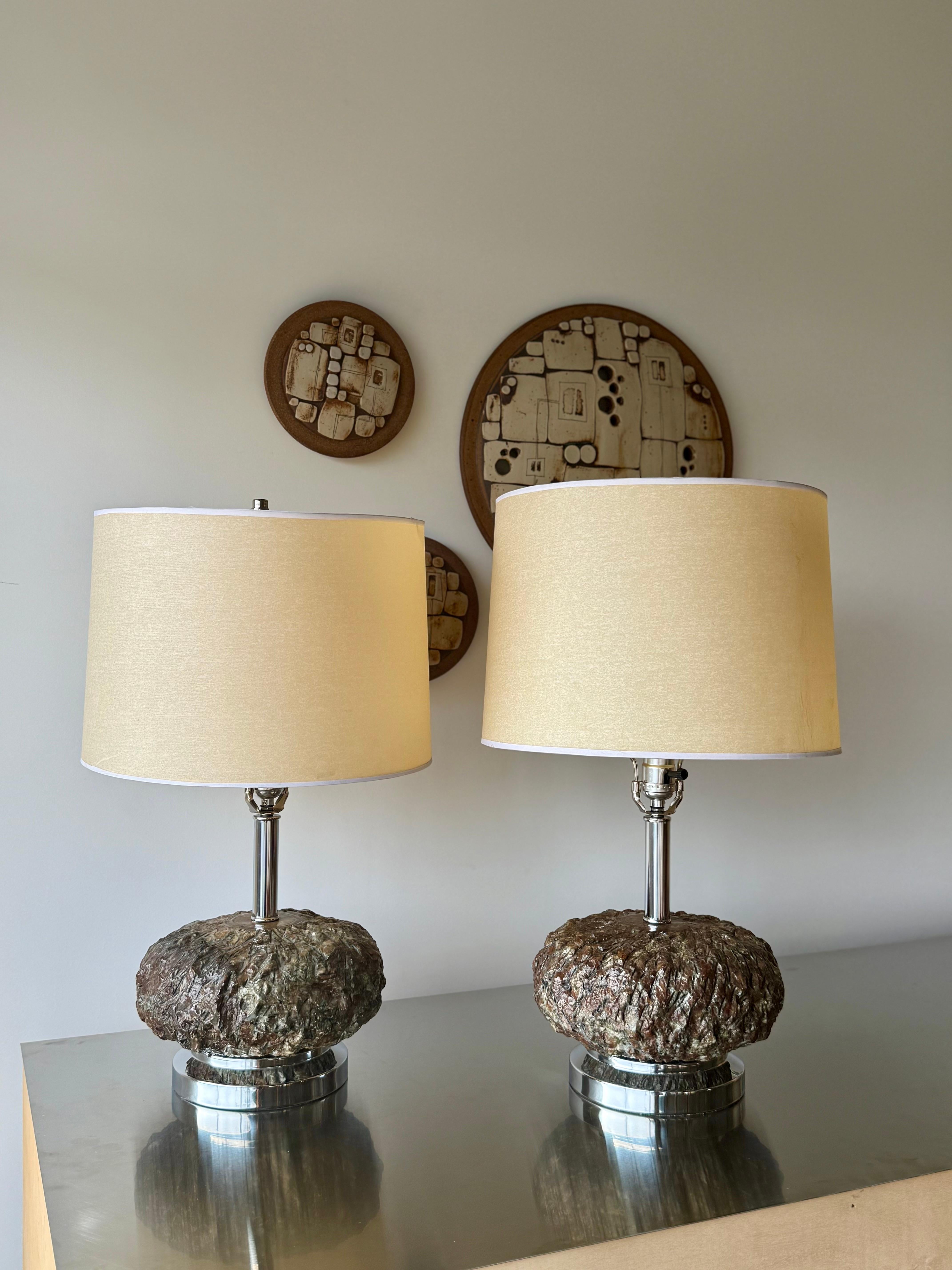 Sculptural Brutalist-style set of table lamps featuring a heavily textured base in dark resin/stone composite, set on a polished chrome plinth with matching chrome hardware. The organic form of the bases —resembling natural rock or bark—contrasts