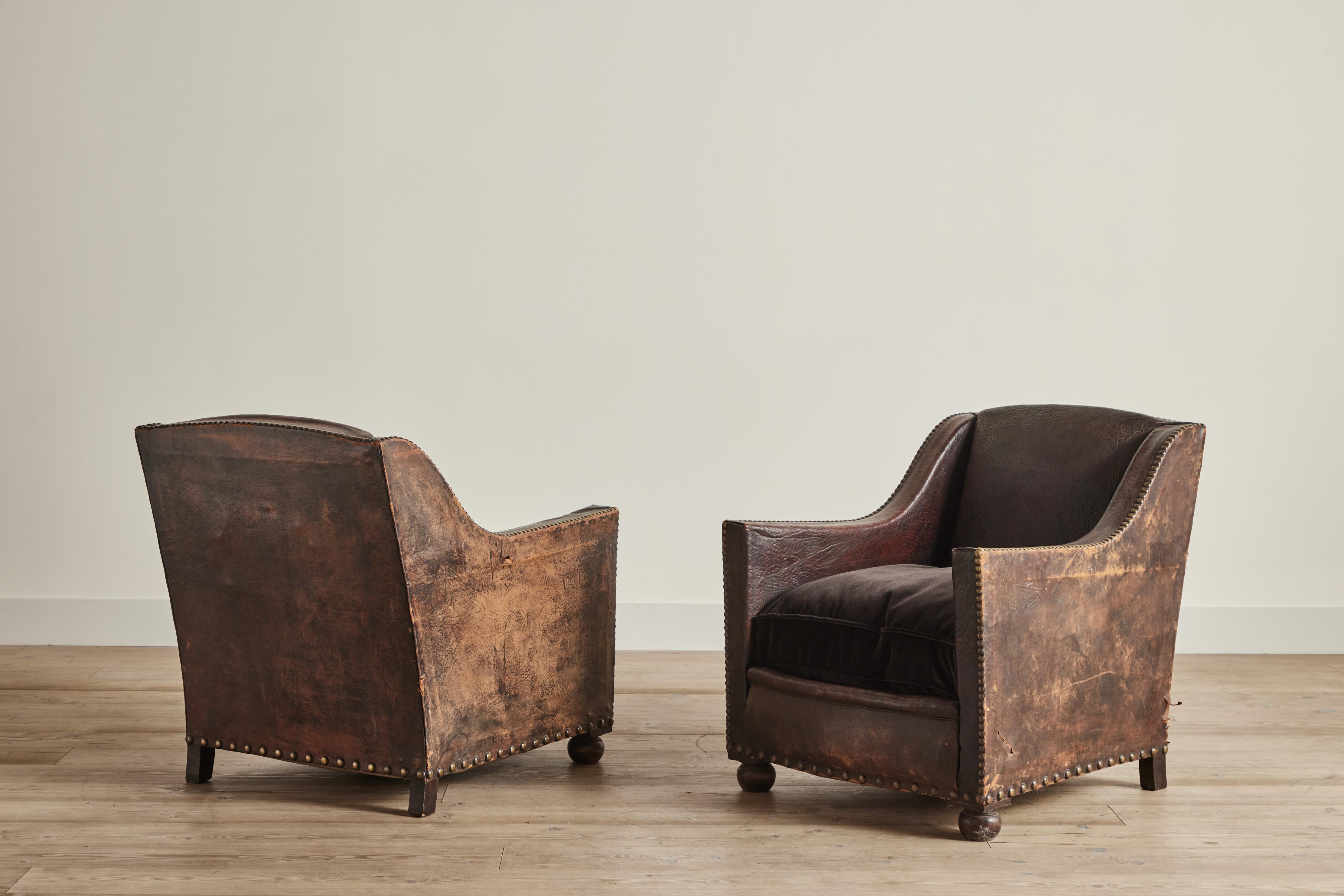 Stunning pair of early 20th century buffalo leather club chairs with brown velvet seat cushions. Chairs sit on wood bun feet and feature brass stud detailing. Visible wear throughout that is consistent with age and use. 