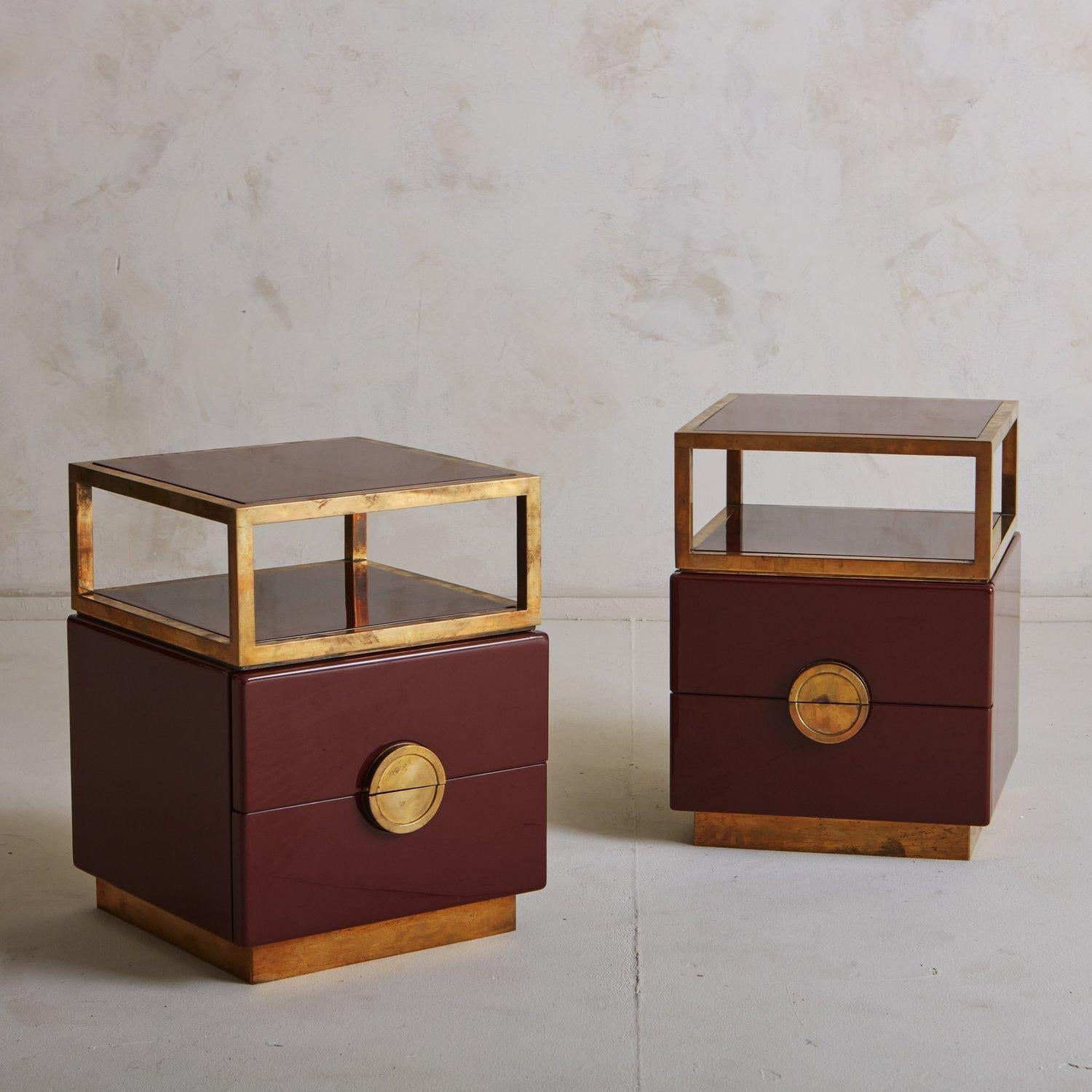 A pair of French end tables featuring a beautiful burgundy lacquered finish with patinated brass detailing and plinth bases. These tables have two drawers with demilune brass pulls and an open upper shelf perfect for styling books and trinkets.