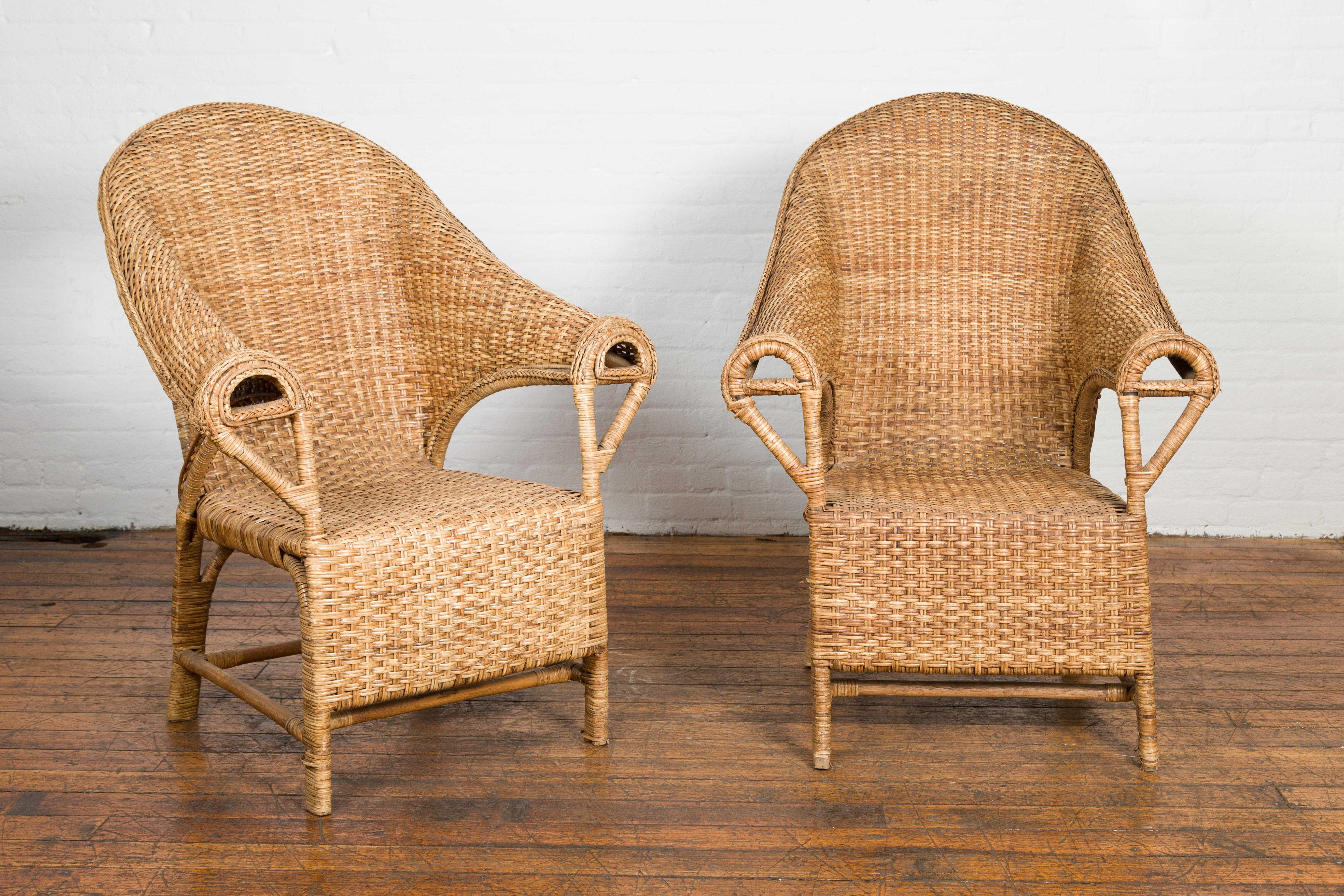A vintage pair of Country style Burmese hand woven rattan lounge chairs from the mid 20th century, with looping, out scrolling arms, arching backs and cross stretchers. Created in Burma (nowadays called Myanmar) during the midcentury period, this